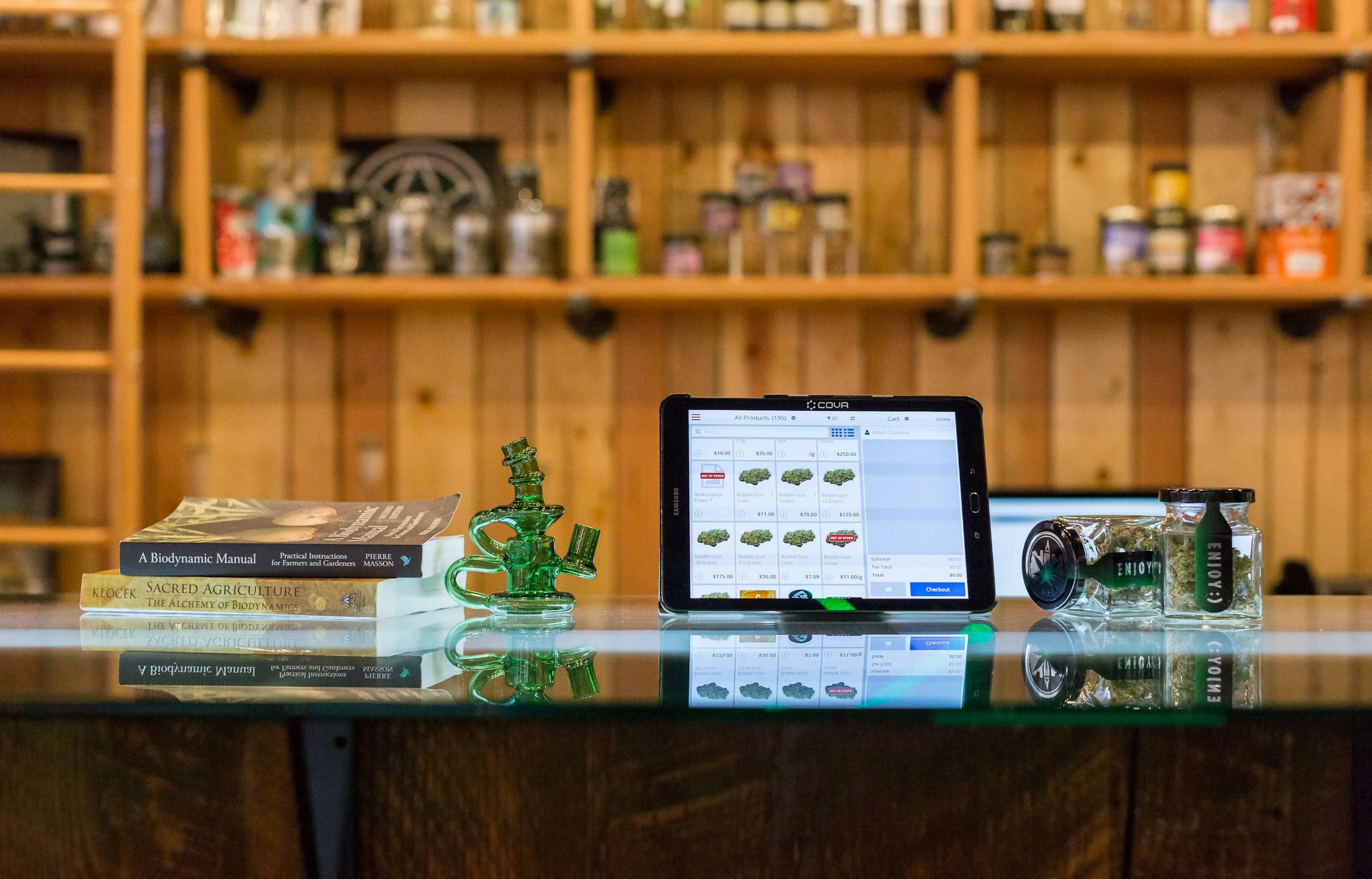 A wooden shelf with various jars and containers, a digital tablet displaying an menu or shopping list, a green glass pipe, and some books about biodynamics and agriculture on a reflective surface.