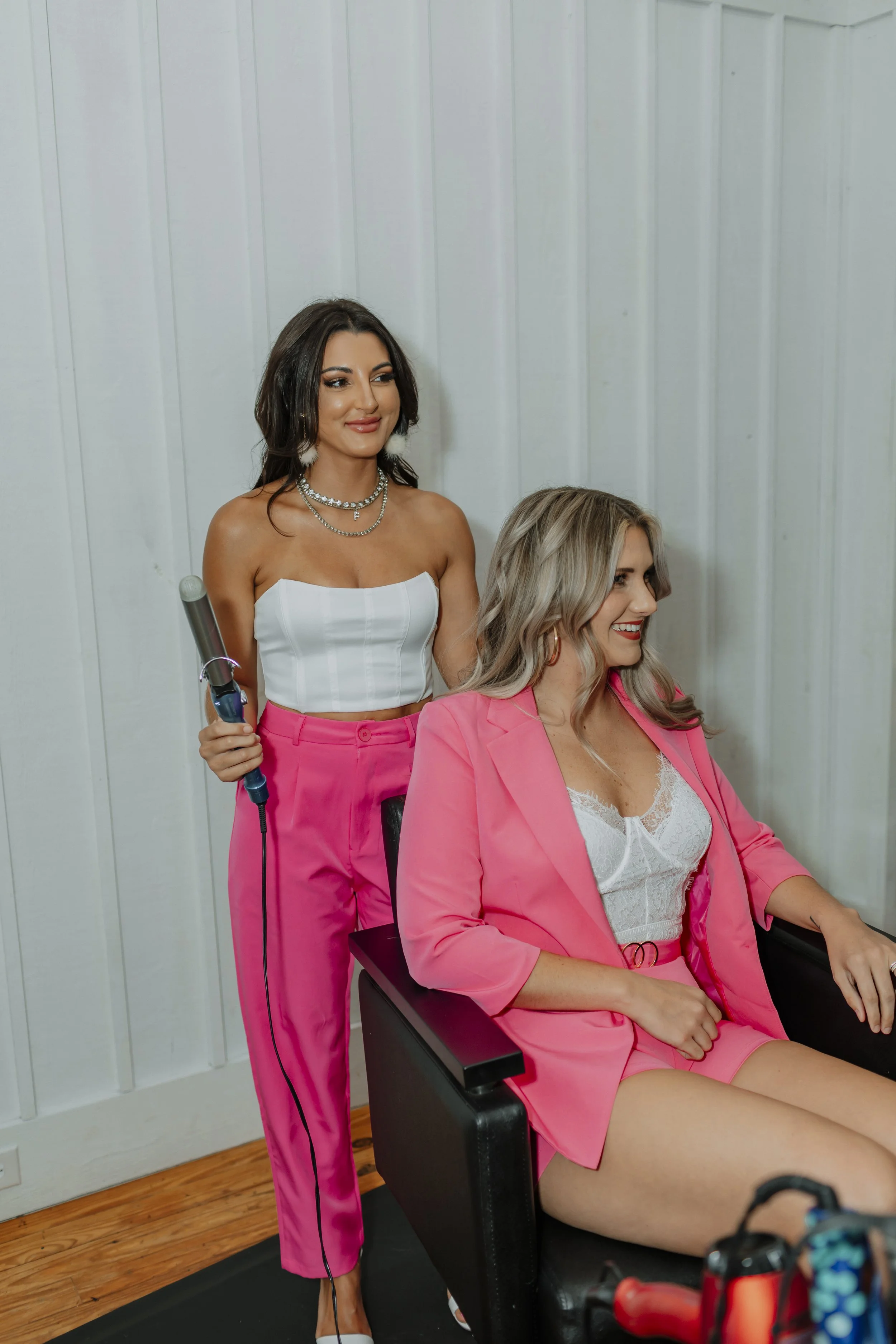 A woman with dark hair wearing a white top and pink pants is styling another woman with blonde hair, who is sitting in a black chair, wearing a light pink blazer and matching shorts, and a white lace top.