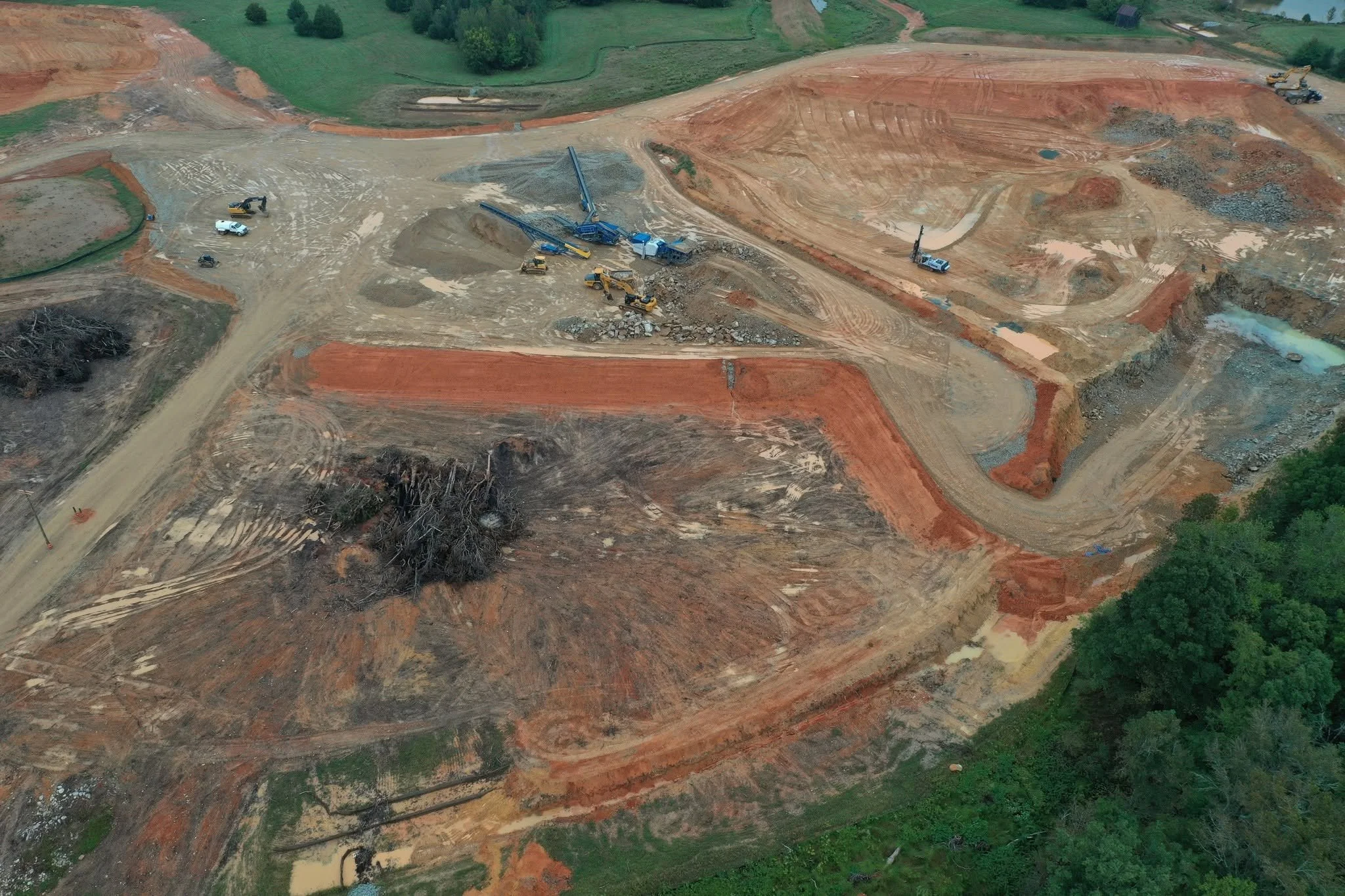An aerial view of a construction site with dirt, construction vehicles, and green trees surrounding the area.