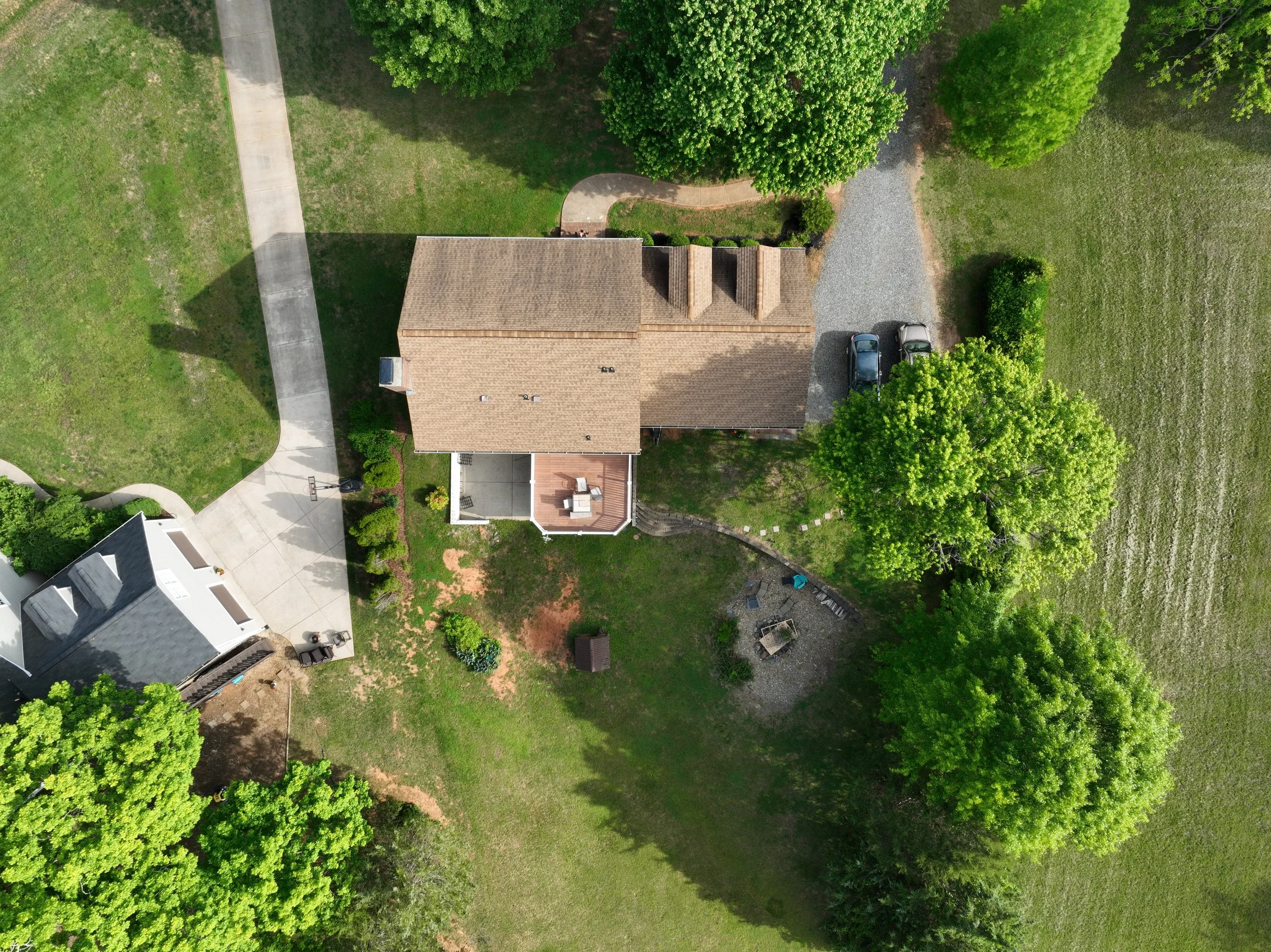 An aerial view of a residential backyard with a house, surrounding greenery, trees, and a driveway with parked cars.