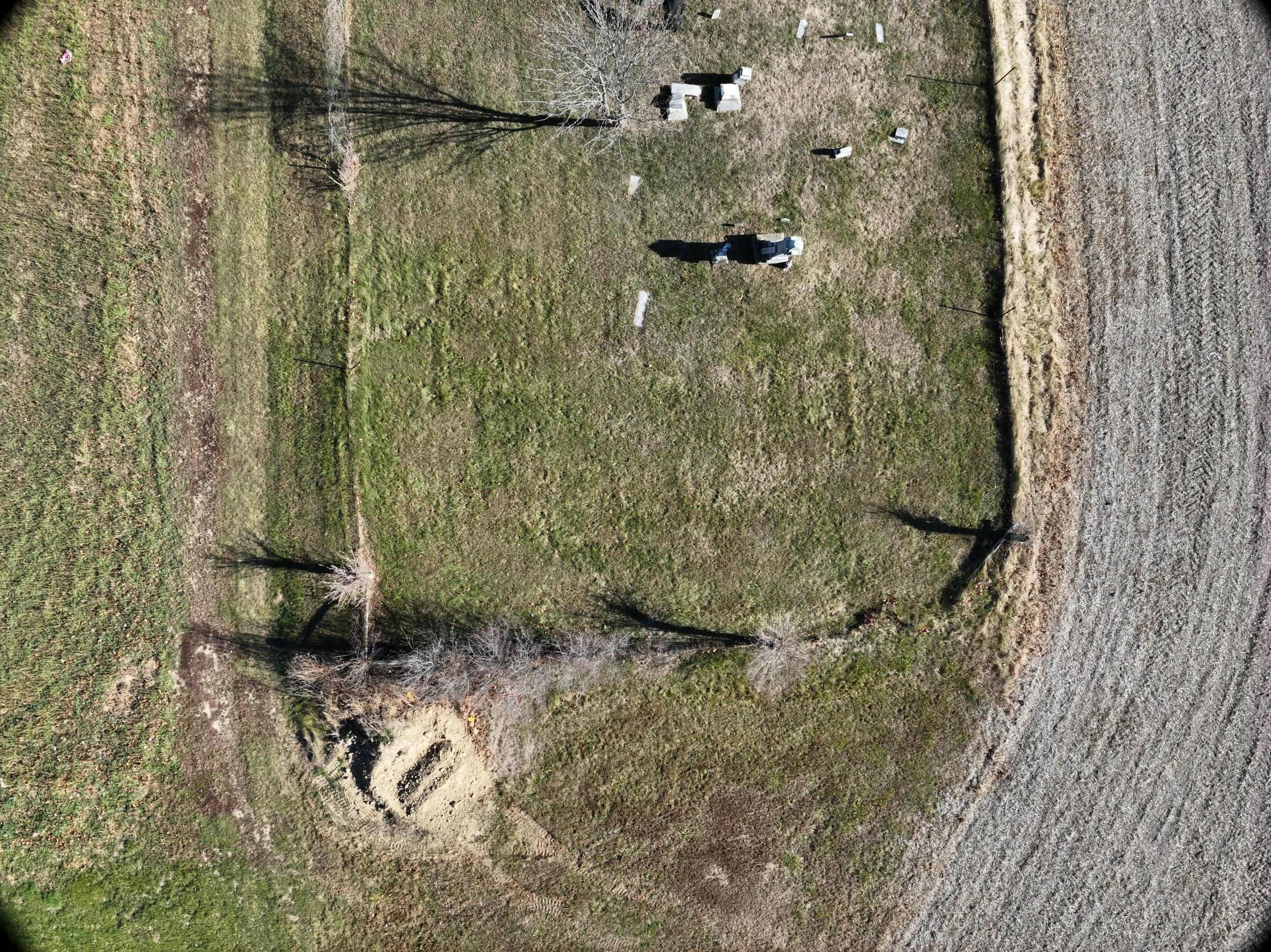 An aerial view of a grassy area with a few surrounding trees, some of which are leafless. There is a small stone structure or monument at the top center of the image. The land appears to have some patches of dirt or wear, particularly around the monument.