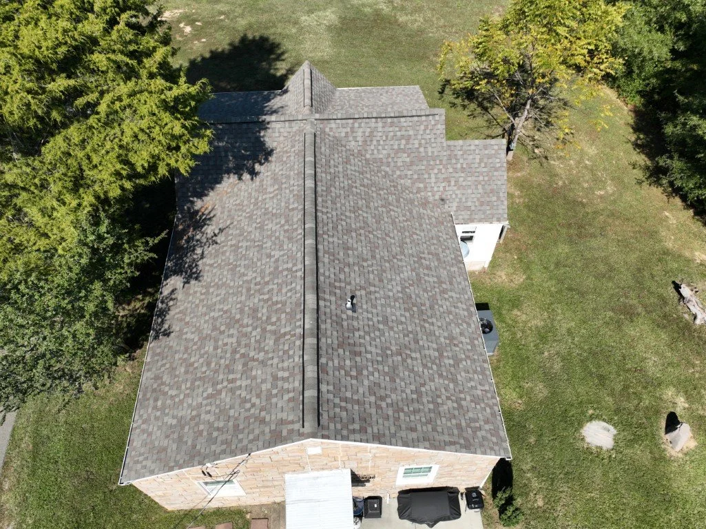 An aerial view of a house with a shingled roof, surrounded by green trees and a grassy yard.