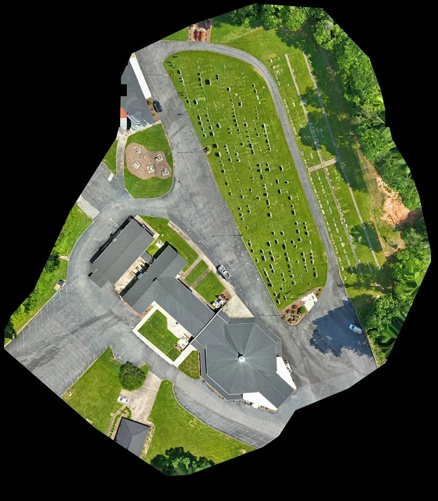 An aerial view of a church with a parking lot, a cemetery with rows of headstones, a playground, and surrounding greenery.