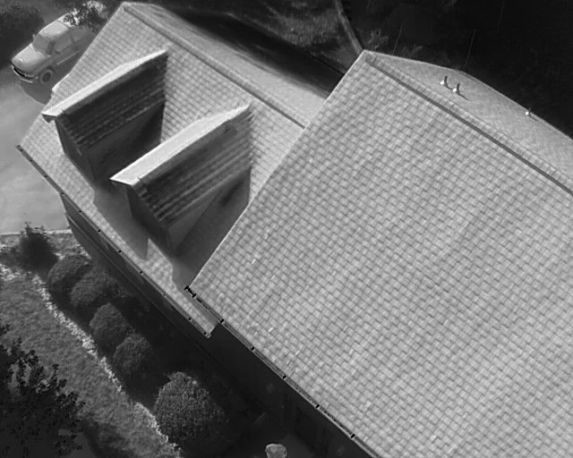 Aerial view of a house with a tiled roof, showing two skylights, surrounded by bushes and parked cars.