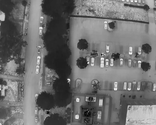 An aerial view of a parking lot with scattered cars, some trees, and a building with rooftop structures.