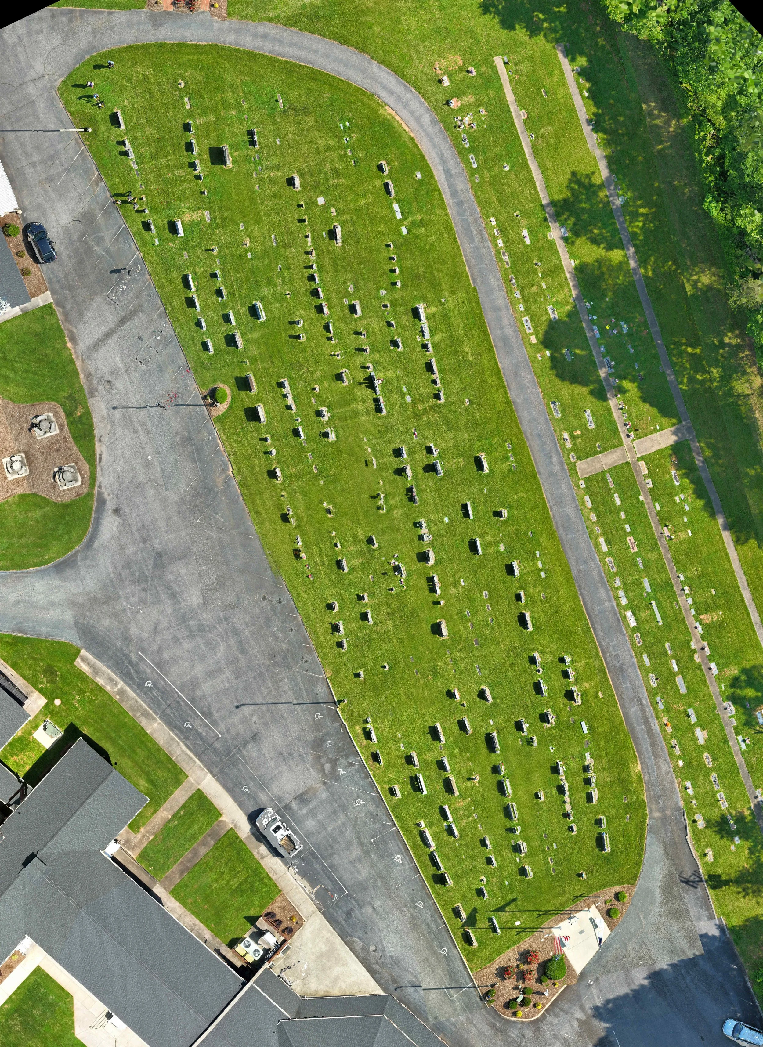 An aerial view of a cemetery with numerous grave markers on a green lawn, surrounded by roads and a parking lot, with trees on the right side.