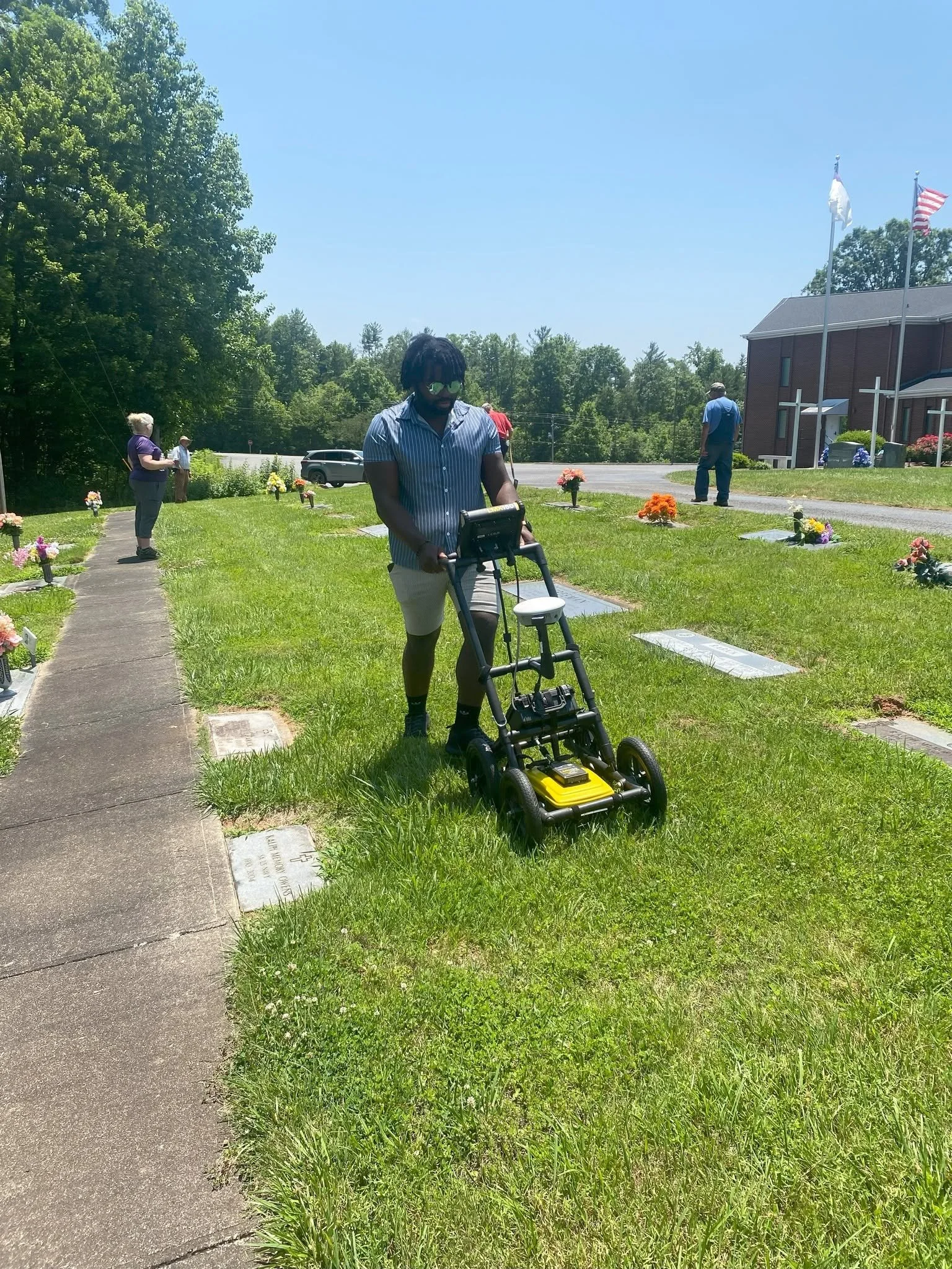 People visiting a cemetery on a sunny day. One man using a walker is in the foreground, and others are standing near graves marked with flowers. Flags are flying in the background near a building.