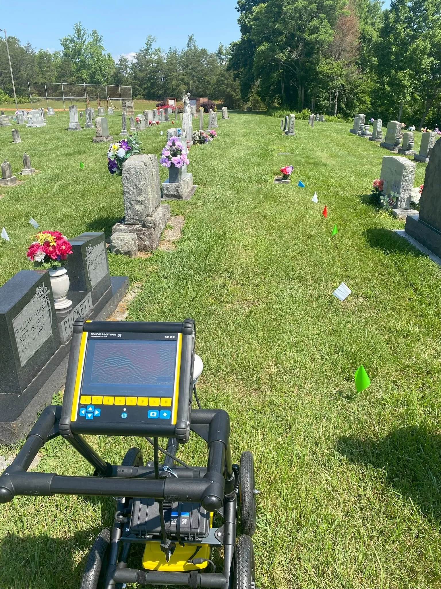 View of a cemetery with headstones and flowers, with ground penetrating radar, tall trees and a blue sky in the background.