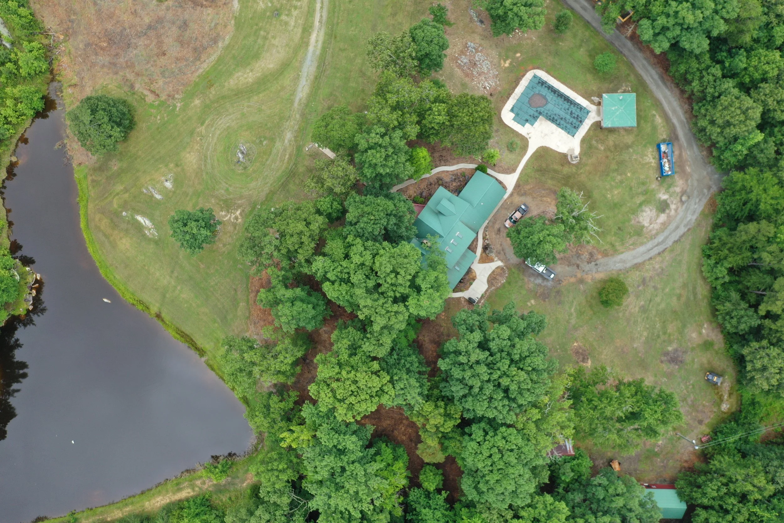 An aerial view of a green yard with trees, a pond, and a building with a green roof. There is also a paved area with a swimming pool and a small shelter nearby, along with a few parked vehicles.