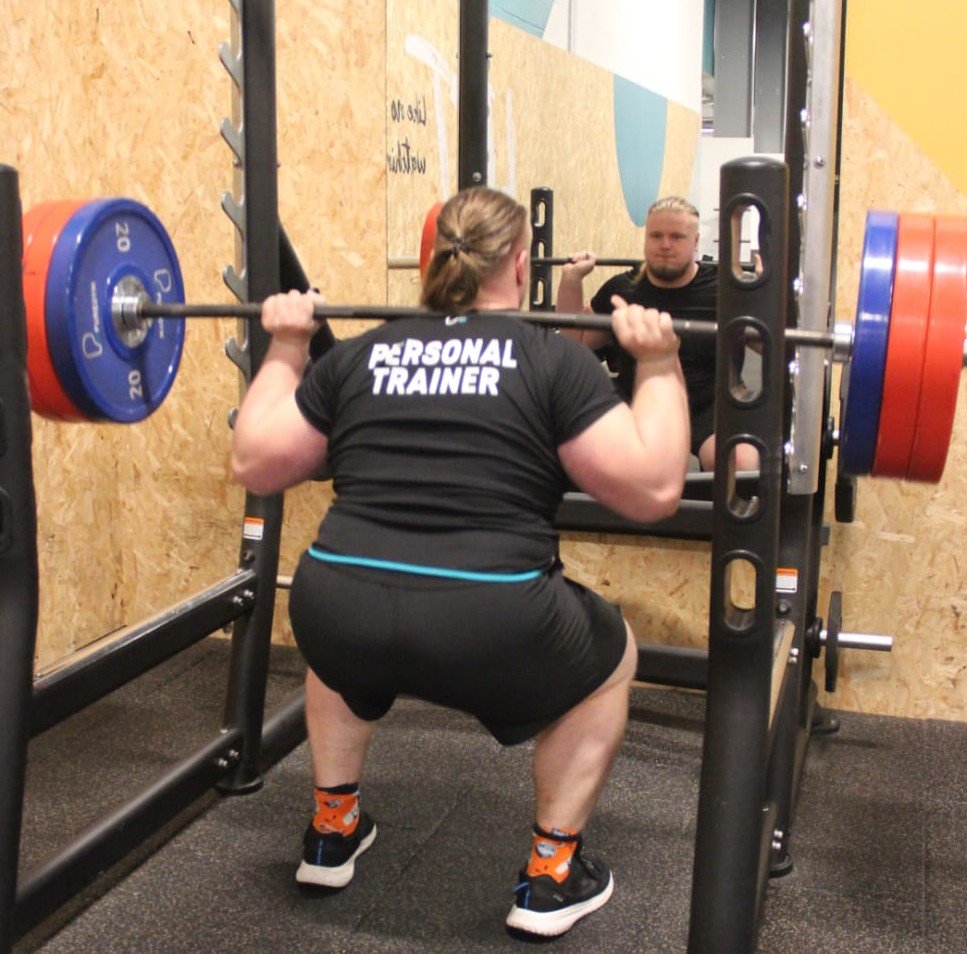 A person performs a squat exercise in a gym, with the word 'PERSONAL TRAINER' printed on the back of their t-shirt, holding a barbell loaded with red and blue weights at shoulder level, in front of a mirror.