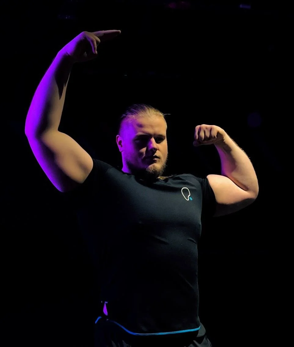 A man with a beard and long hair pulled back, flexing his biceps and pointing upward with his left hand, standing in front of a dark background with purple lighting.