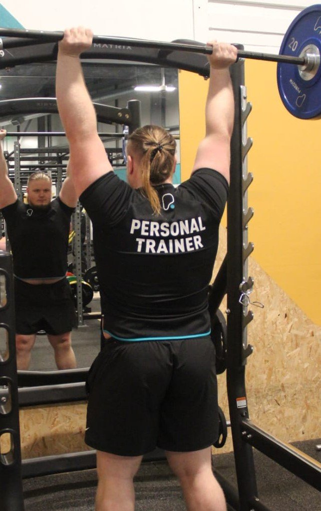 A person with their back to the camera lifting a barbell in a gym. They are wearing a black shirt with 'PERSONAL TRAINER' written on the back. There is a mirror reflecting their face and part of the gym equipment.