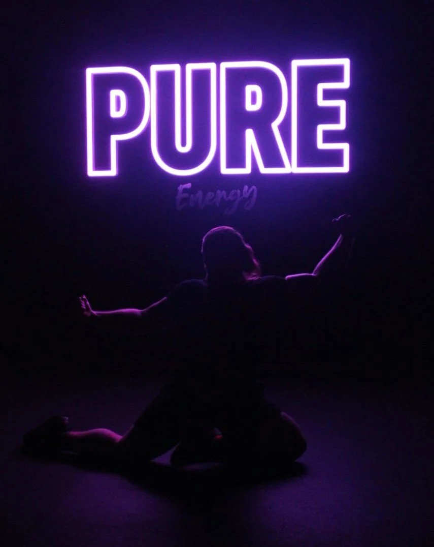 Silhouette of a person kneeling with arms outstretched in front of a neon sign that reads 'PURE' and 'Energy' in purple.