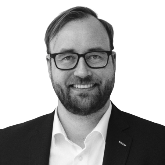 Black and white portrait of a man with glasses, a beard, and a mustache, wearing a suit and white shirt, smiling.