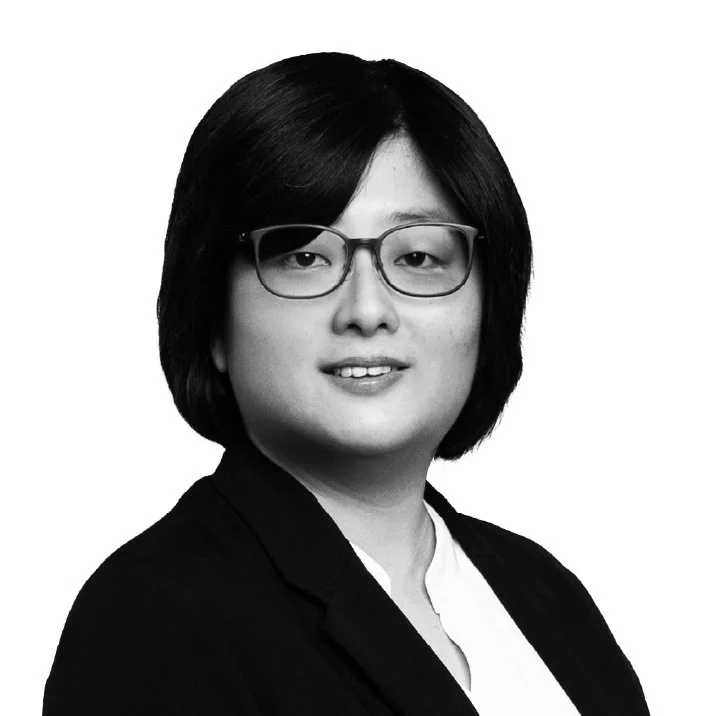 A black-and-white portrait of a woman wearing glasses, with shoulder-length dark hair, a blazer, and a white shirt, looking at the camera against a plain light background.
