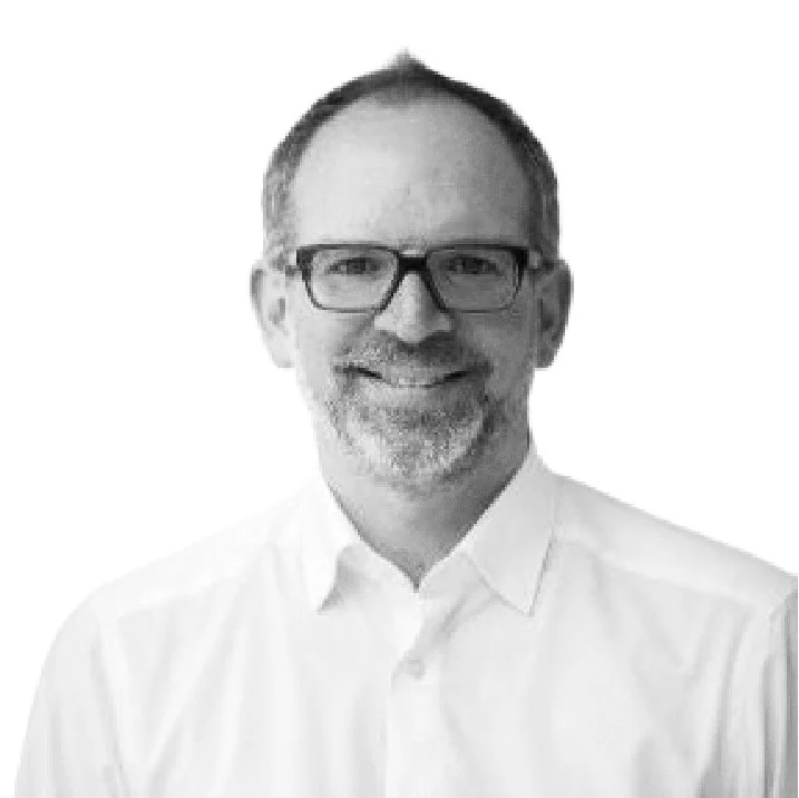A grayscale headshot of a middle-aged man with short hair, glasses, a beard, and a mustache, smiling and wearing a white collared shirt.