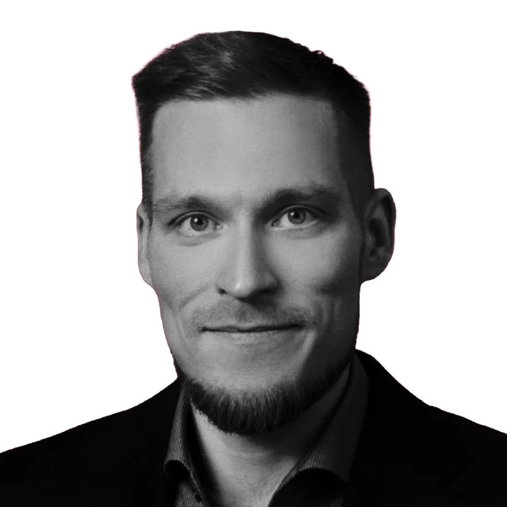 Black and white portrait of a man with short dark hair, a beard, wearing a dark blazer and shirt, smiling slightly, against a plain white background.