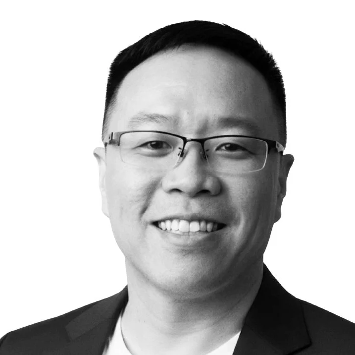 Black and white portrait of a smiling Asian man wearing glasses and a suit.