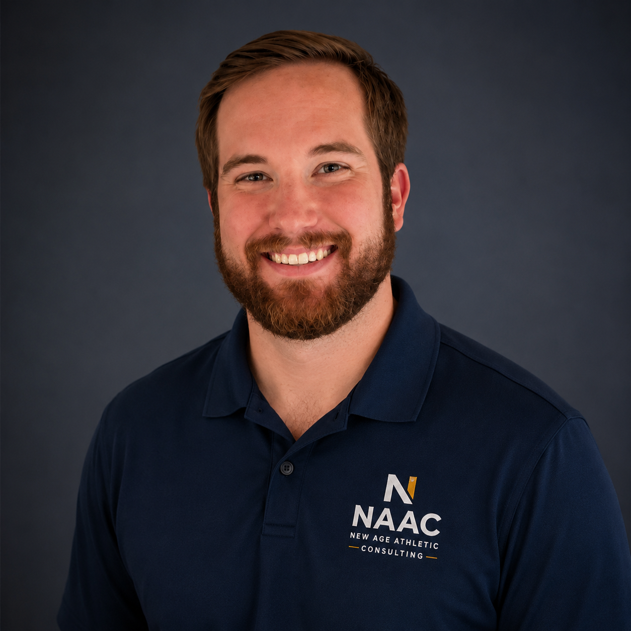 Headshot of a smiling man with a beard and short brown hair wearing a navy blue polo shirt with the NAAC logo and text 'New Age Athletic Consulting' on it, against a dark gray background.
