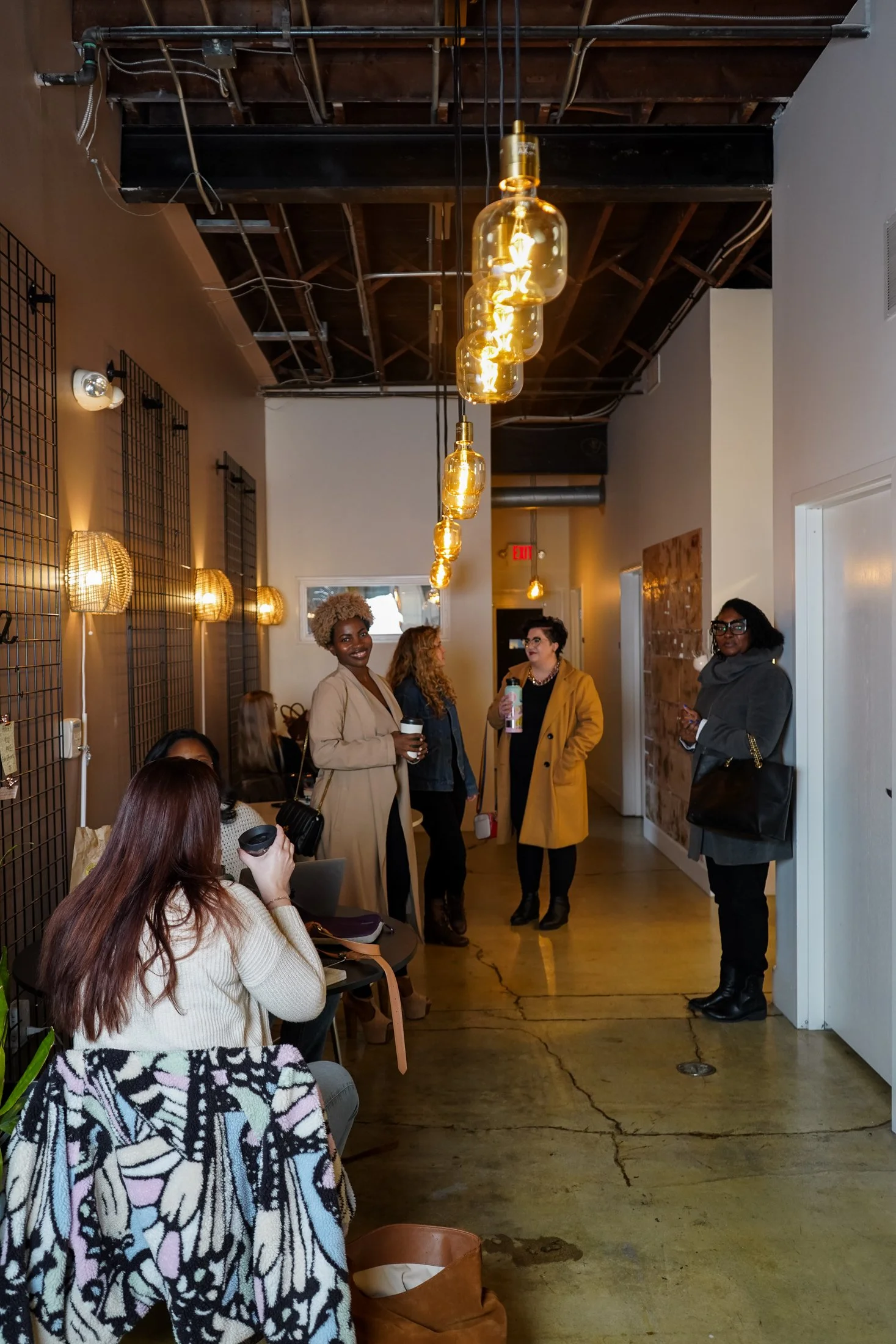 Group of women talking and sitting in a cafe or art gallery with modern lighting and walls, some are holding coffee and wearing stylish coats and sweaters.