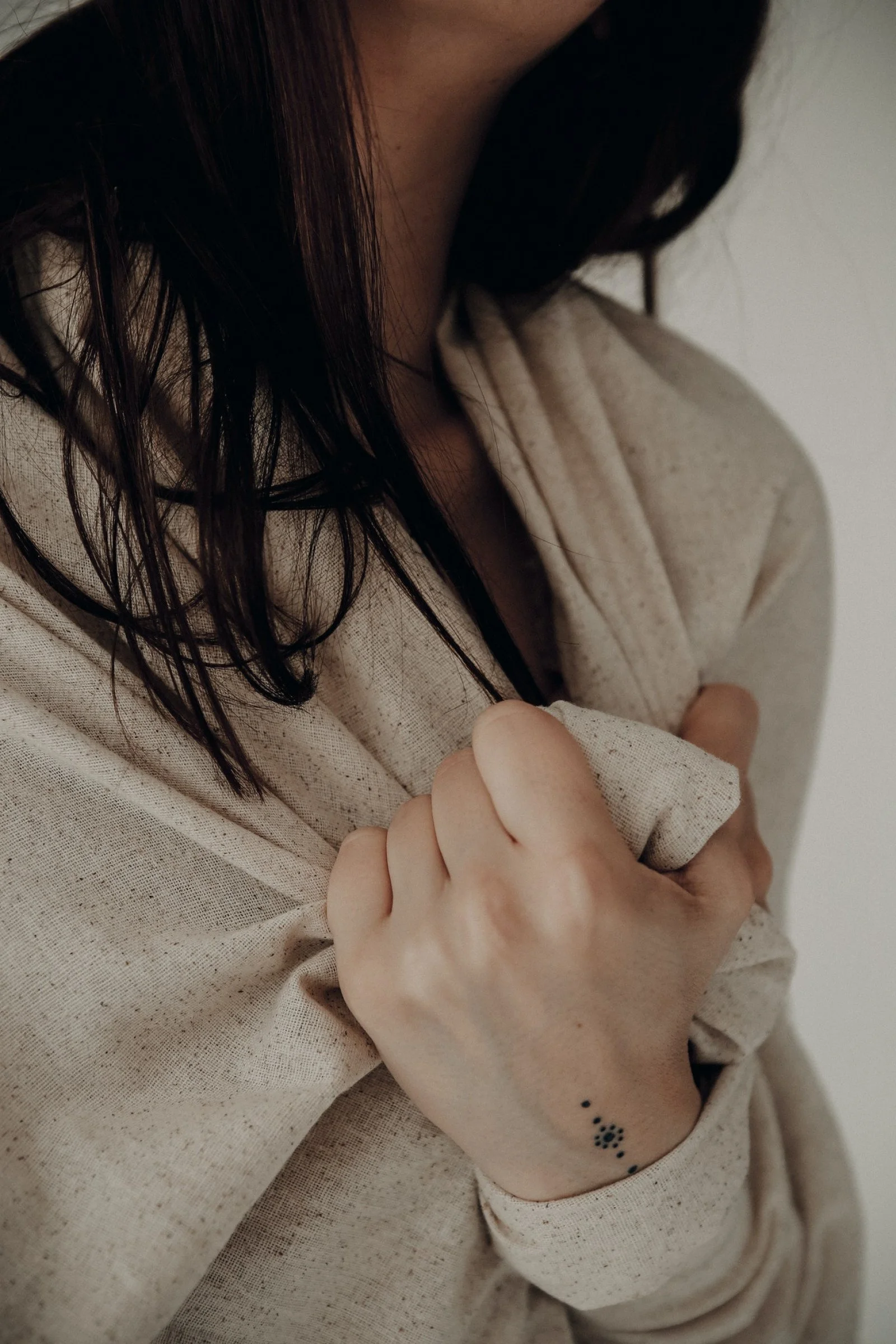 A person with dark hair and light skin wearing a beige hoodie, crossing their arms, exposing a tattoo on their wrist and a small tattoo on their fingers.