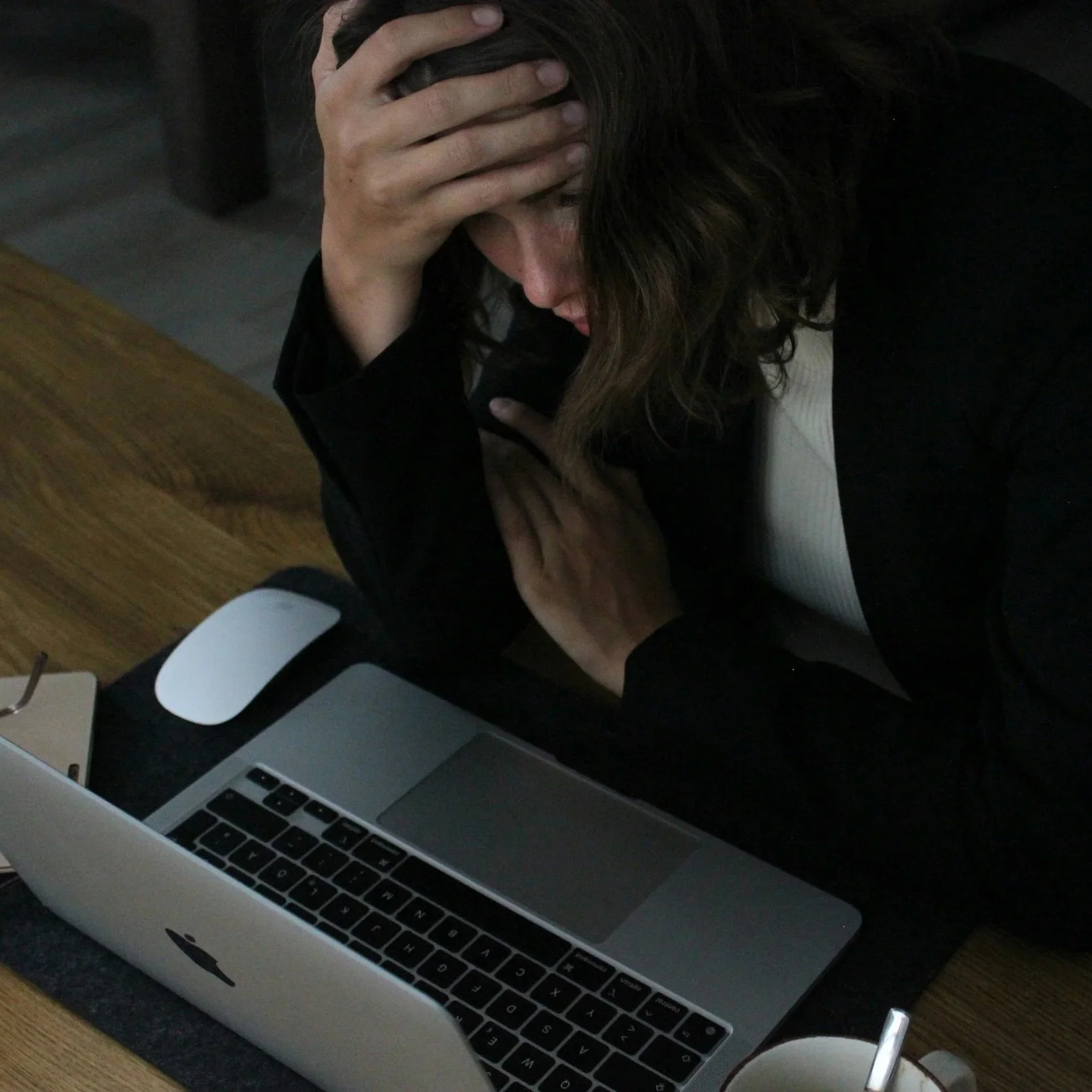 Person sitting at desk with hand on head, laptop open on desk, and a computer mouse nearby, indicating stress or frustration.