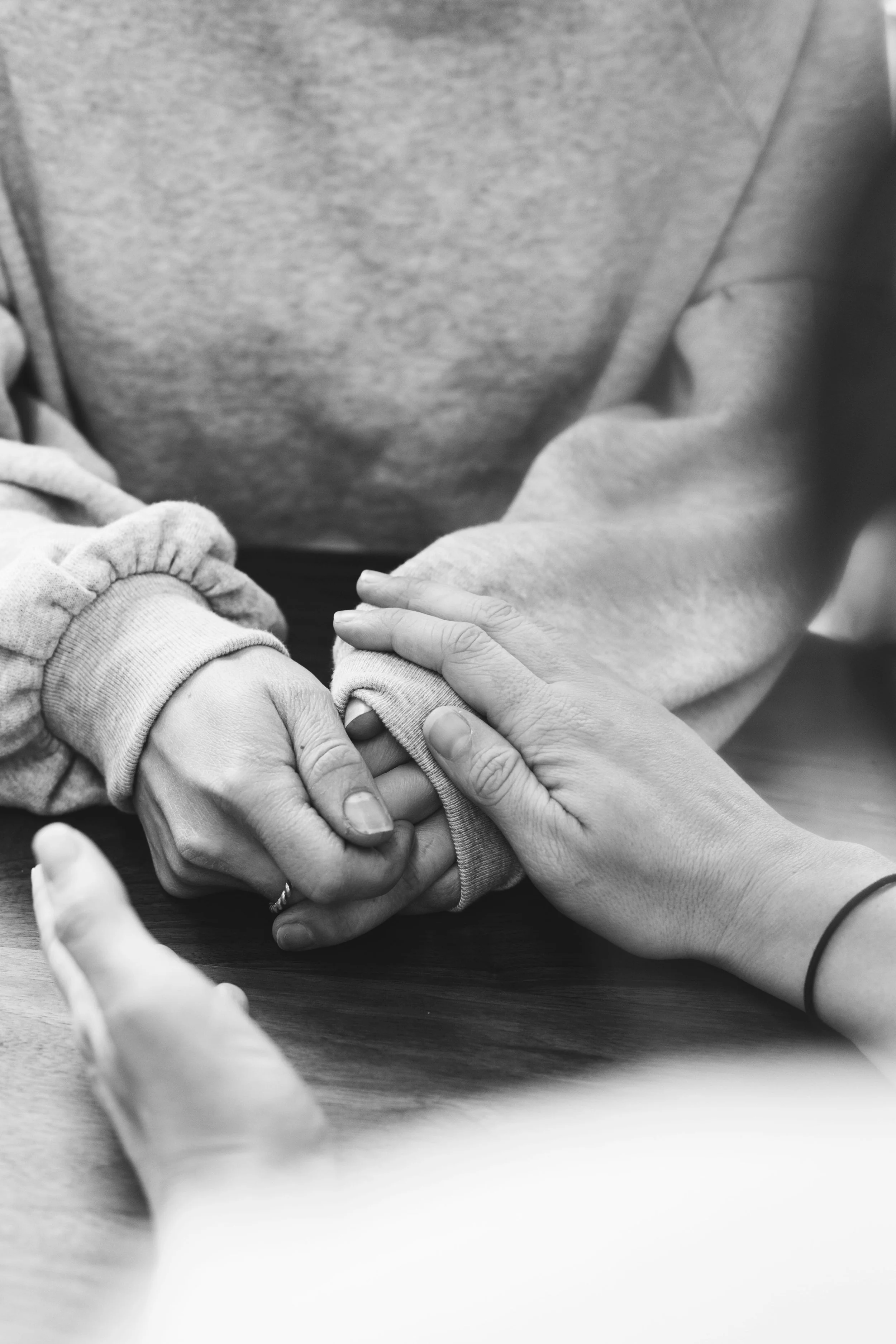 Multiple hands holding and supporting a small child's hand on a table in black and white.
