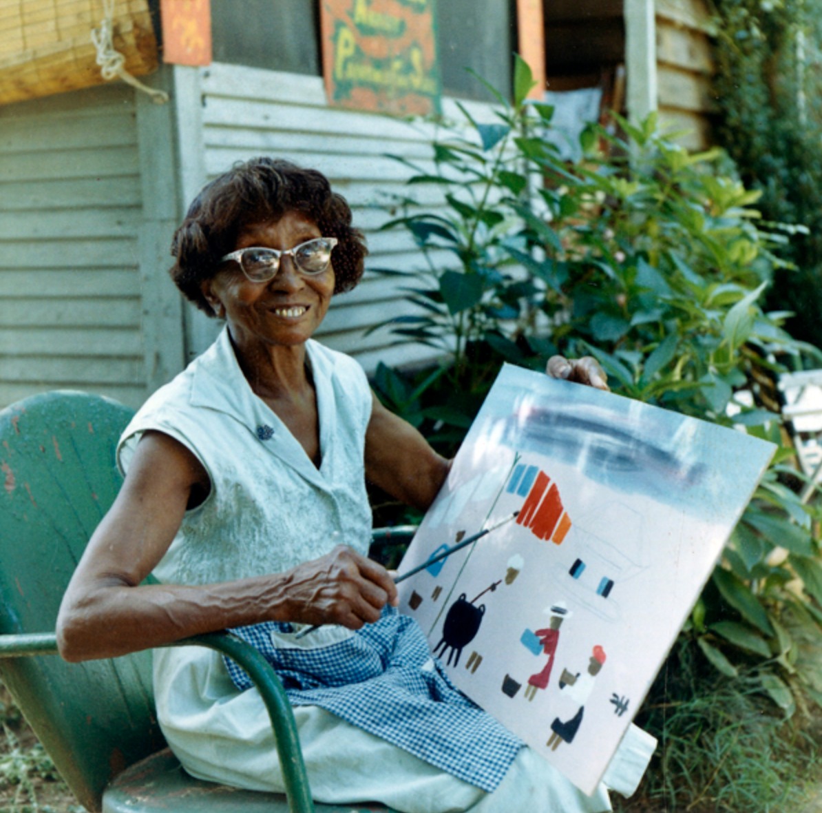 Artist Clementine Hunter sitting in front of her house, smiling at the camera while she holds a painting she is working on.
