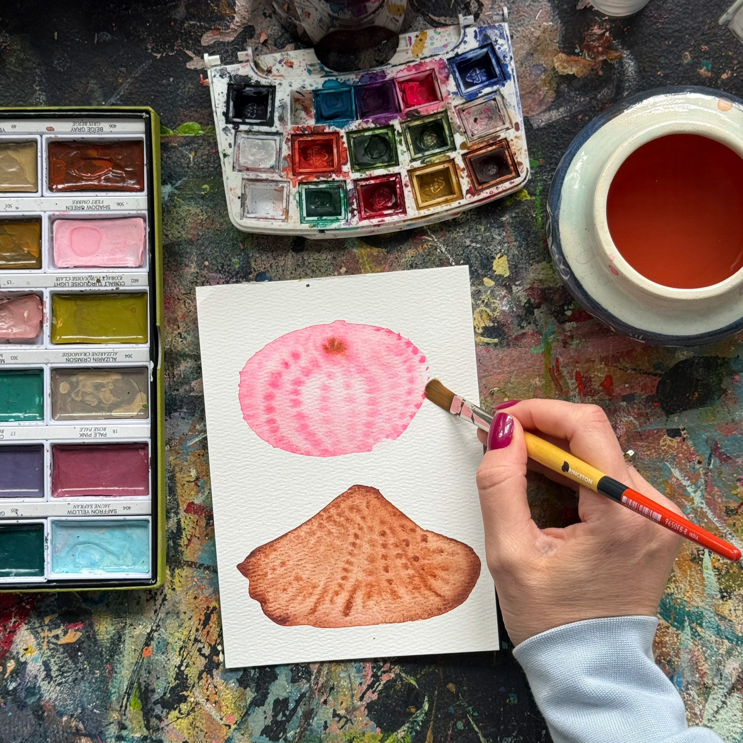 A picture of an artist's desk with watercolors, the artist is holding a brush and painting watercolor seashells.