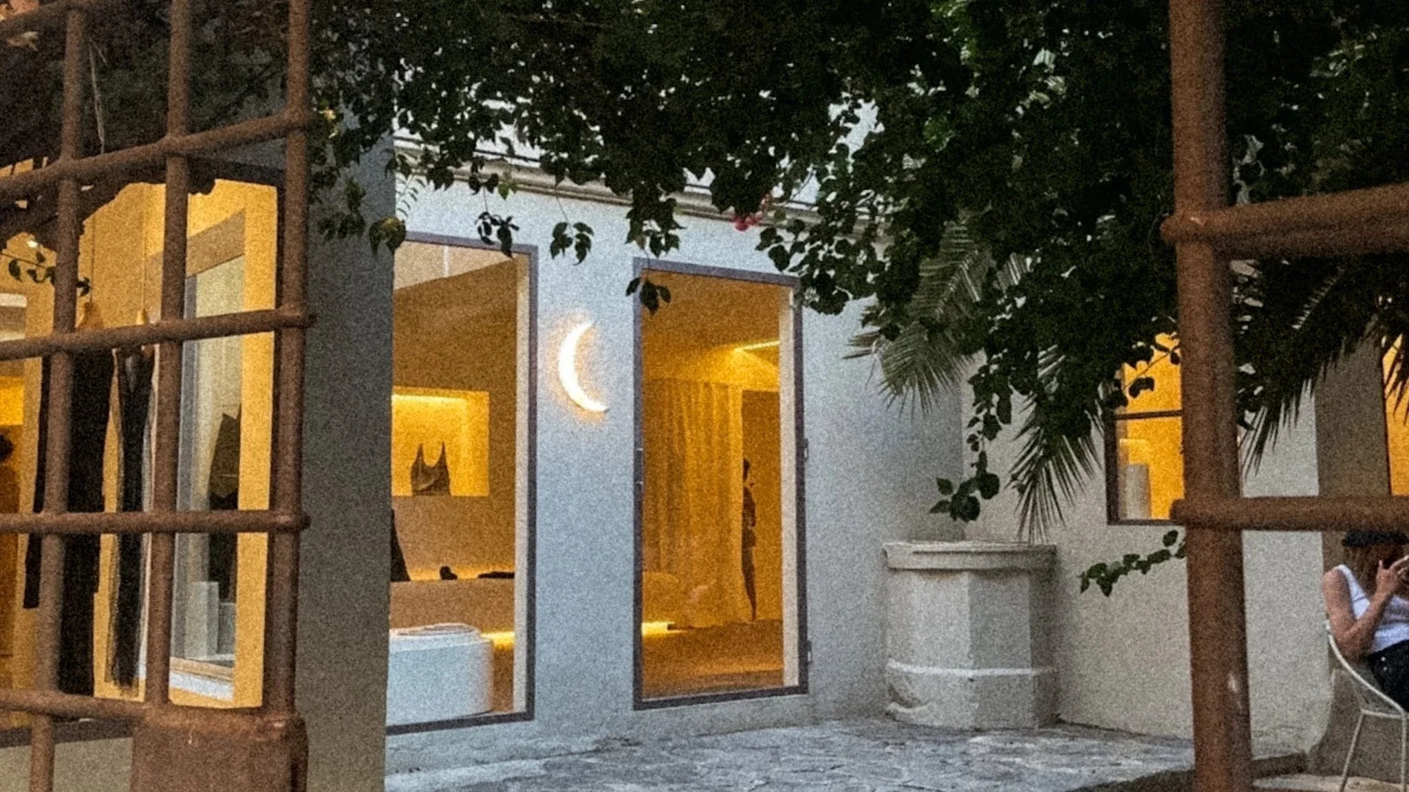 A cobblestone courtyard shaded by large palm trees and fig trees. There is a boutique inside with an entrance marked by a moon-shaped light.