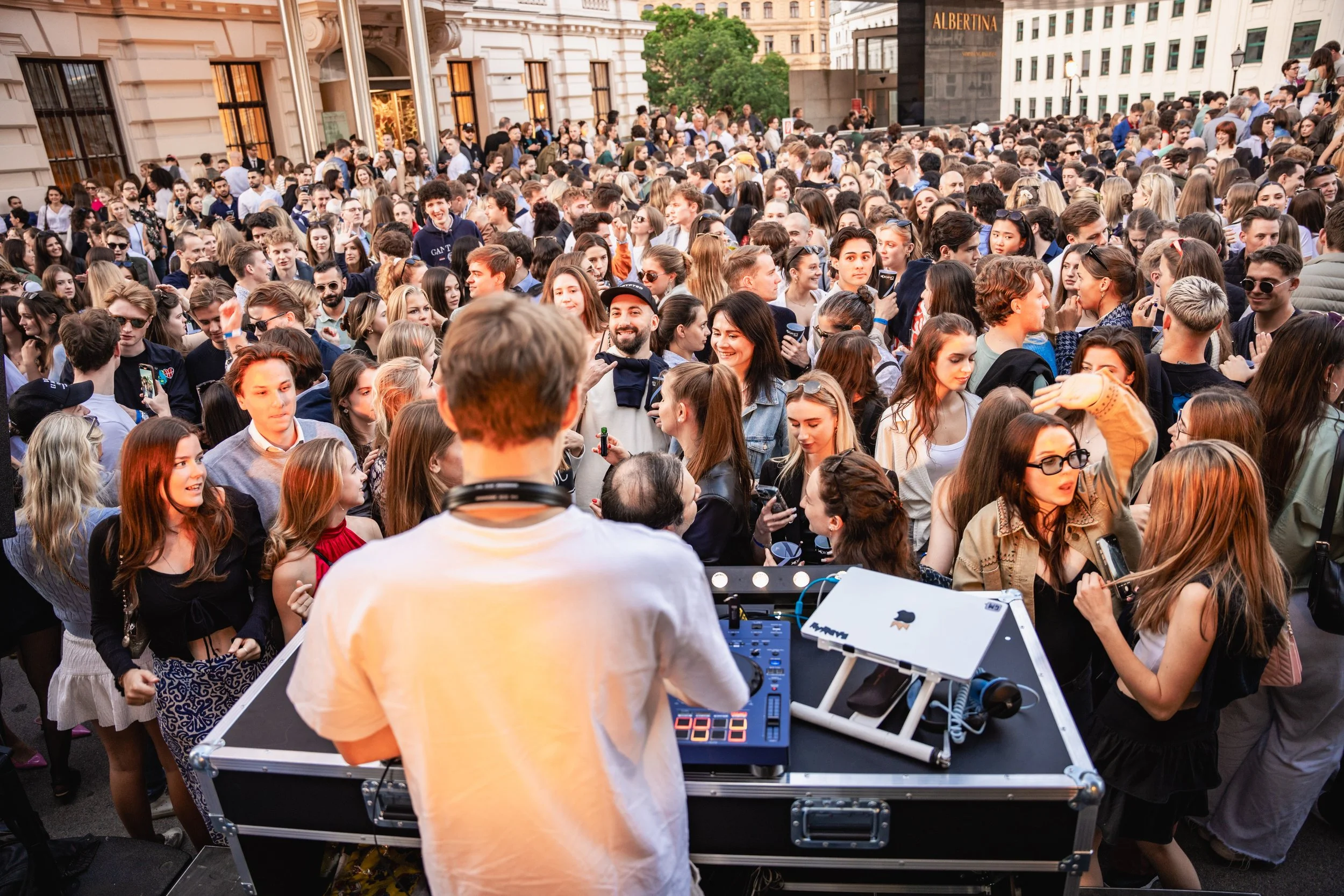 A large crowd of people gathered outdoors in front of historic buildings, some taking photos or videos, with a DJ or music setup in the foreground.