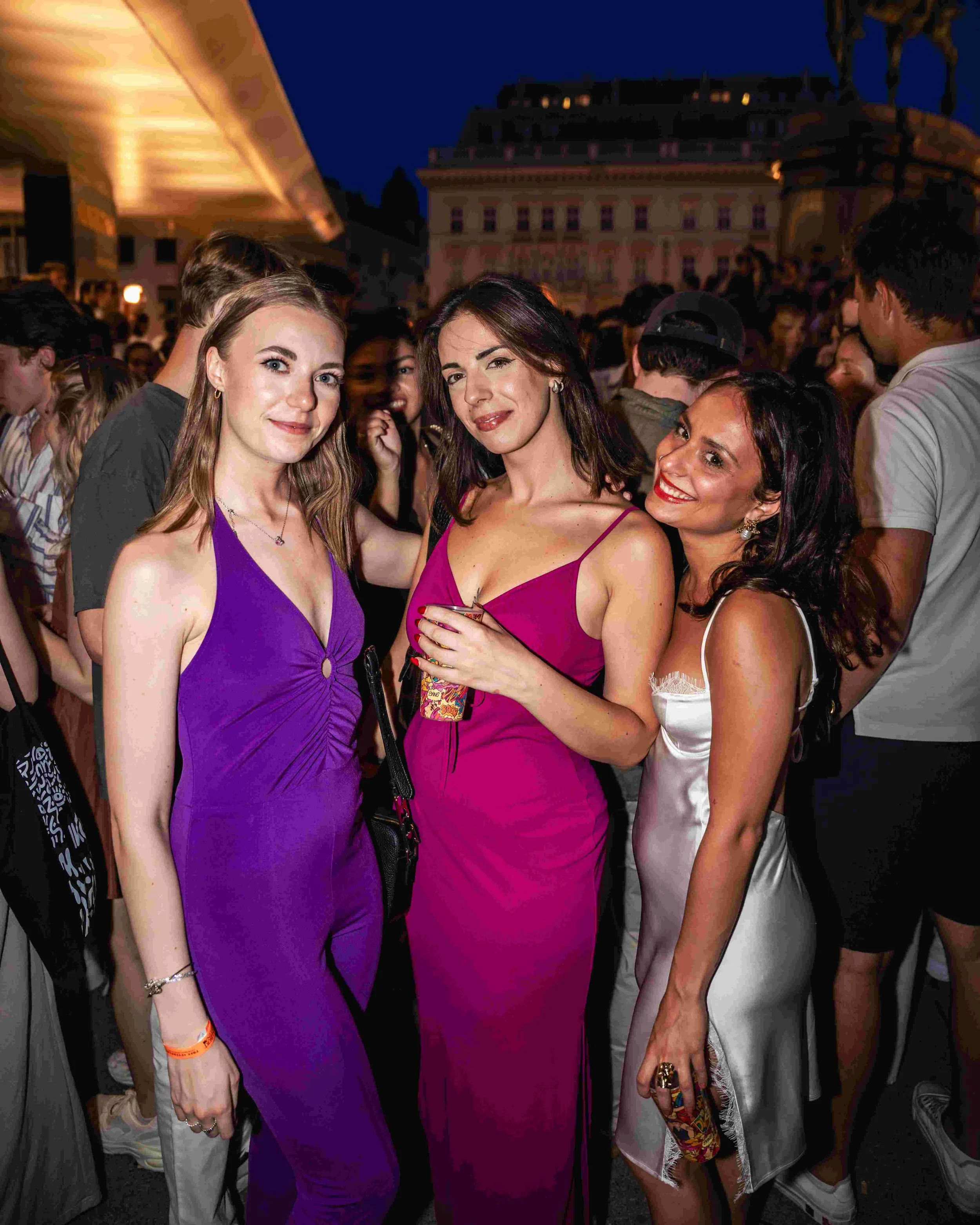 A group of people at a crowded outdoor event at night, with three women in the foreground smiling at the camera. Two women are in purple and pink dresses, and the third woman wears a satin white dress. The background shows other attendees and histori