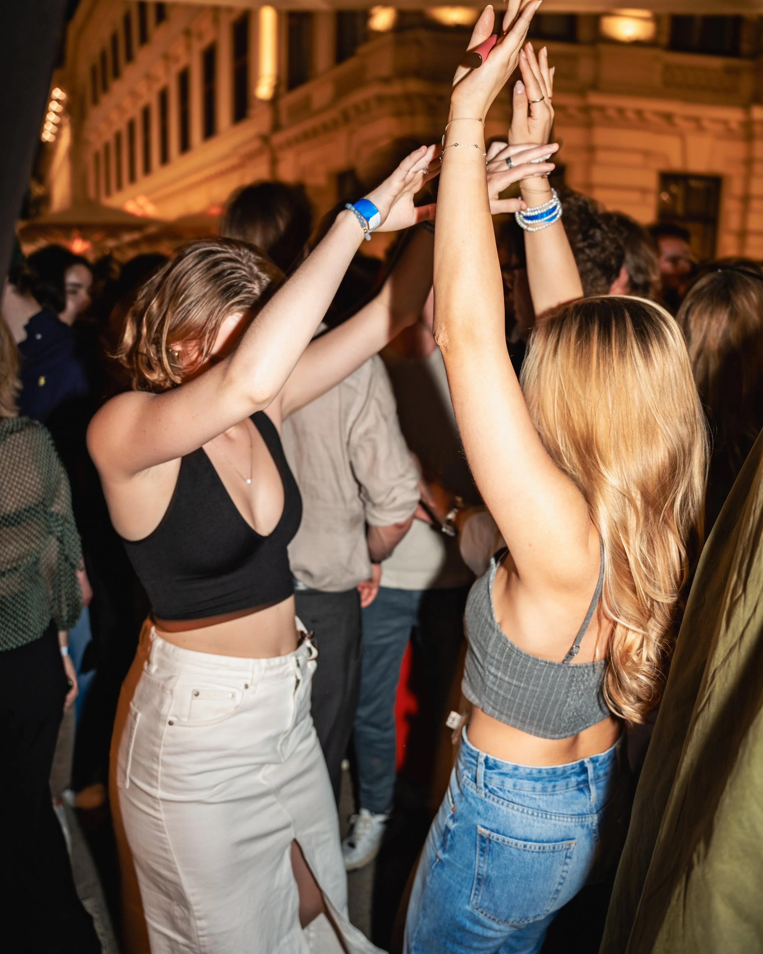 People dancing at a party or nightclub with their hands in the air, in front of a historical building illuminated at night.