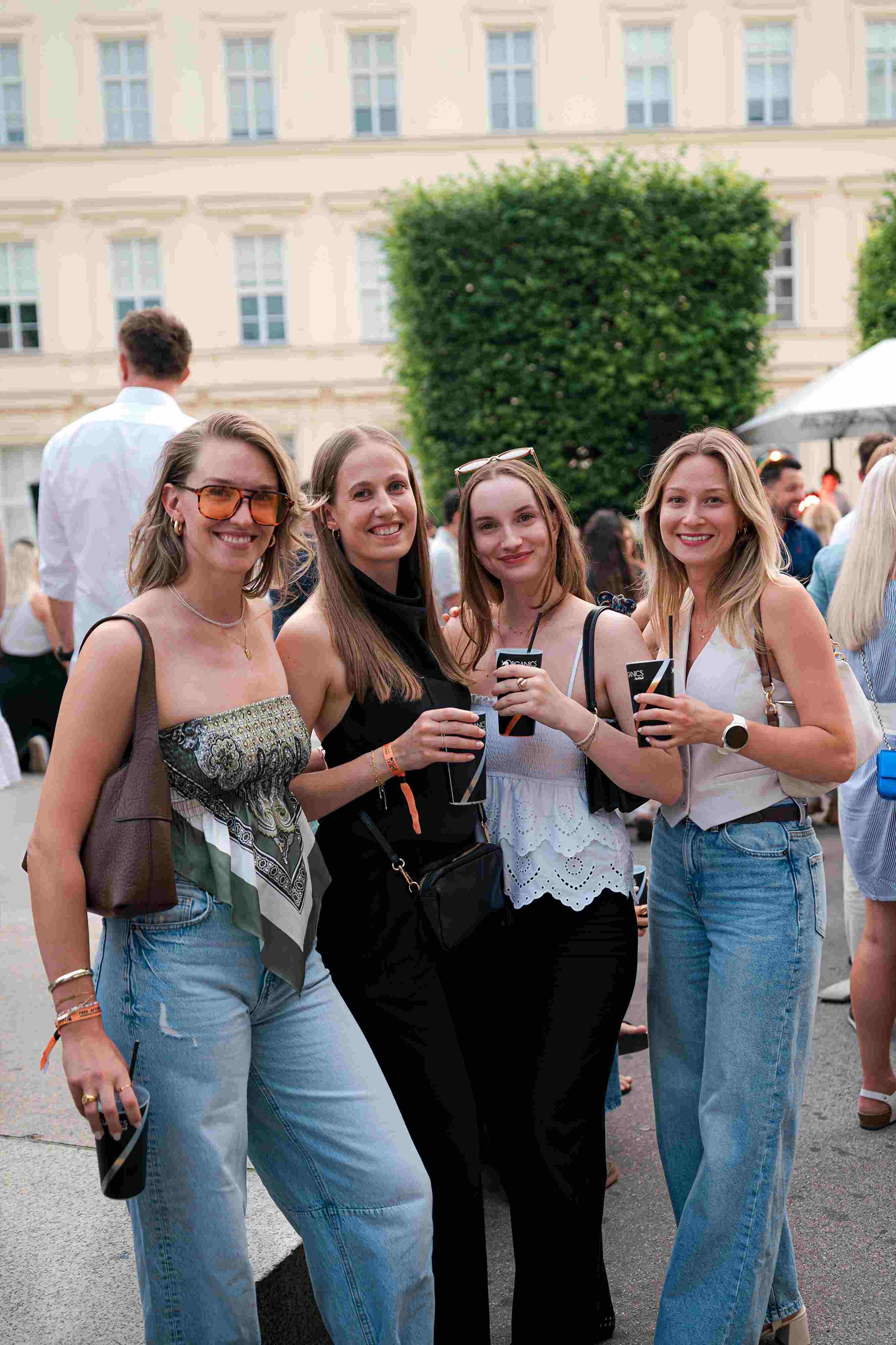 Four women at an outdoor event smiling for the camera, wearing casual summer clothing, with a crowd and a large building in the background.