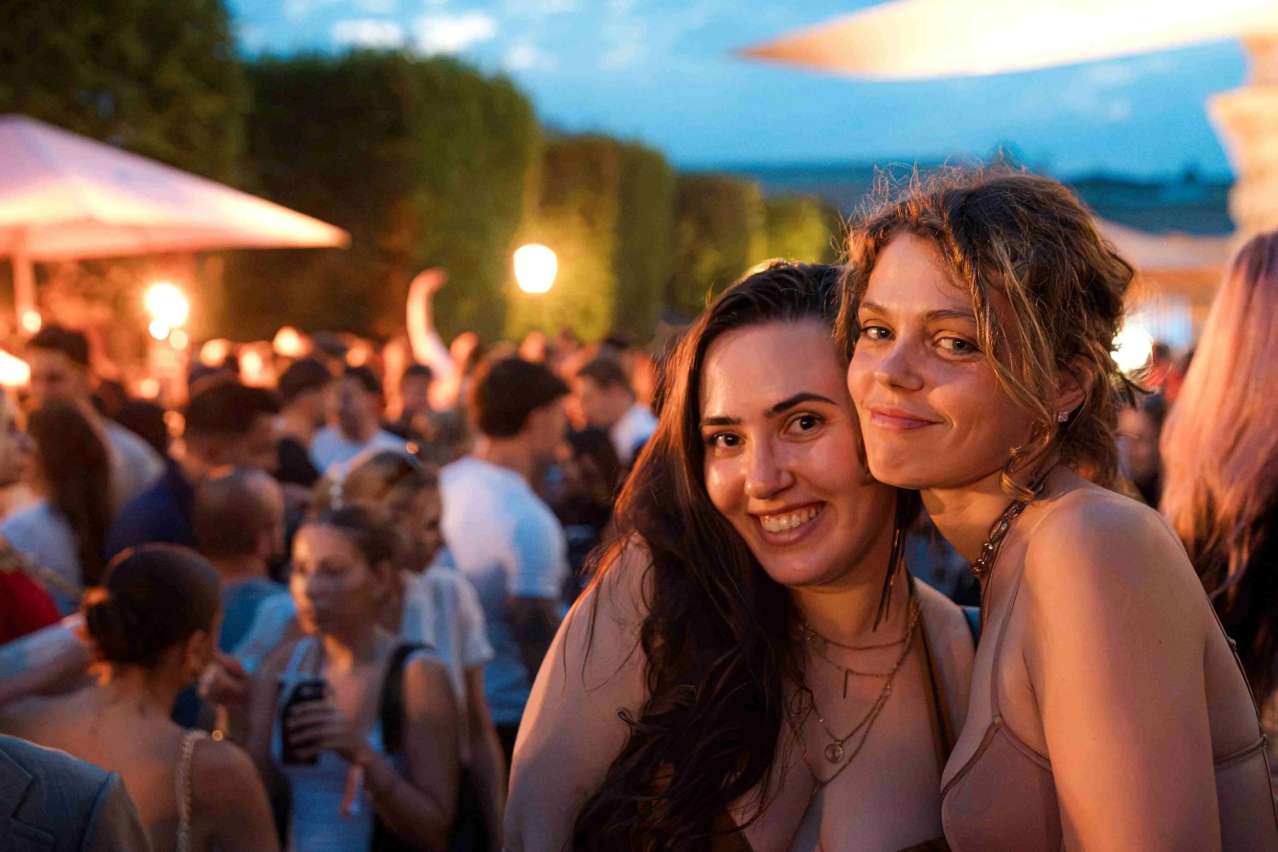 Two women smiling and hugging at an outdoor gathering during sunset with a large crowd in the background.