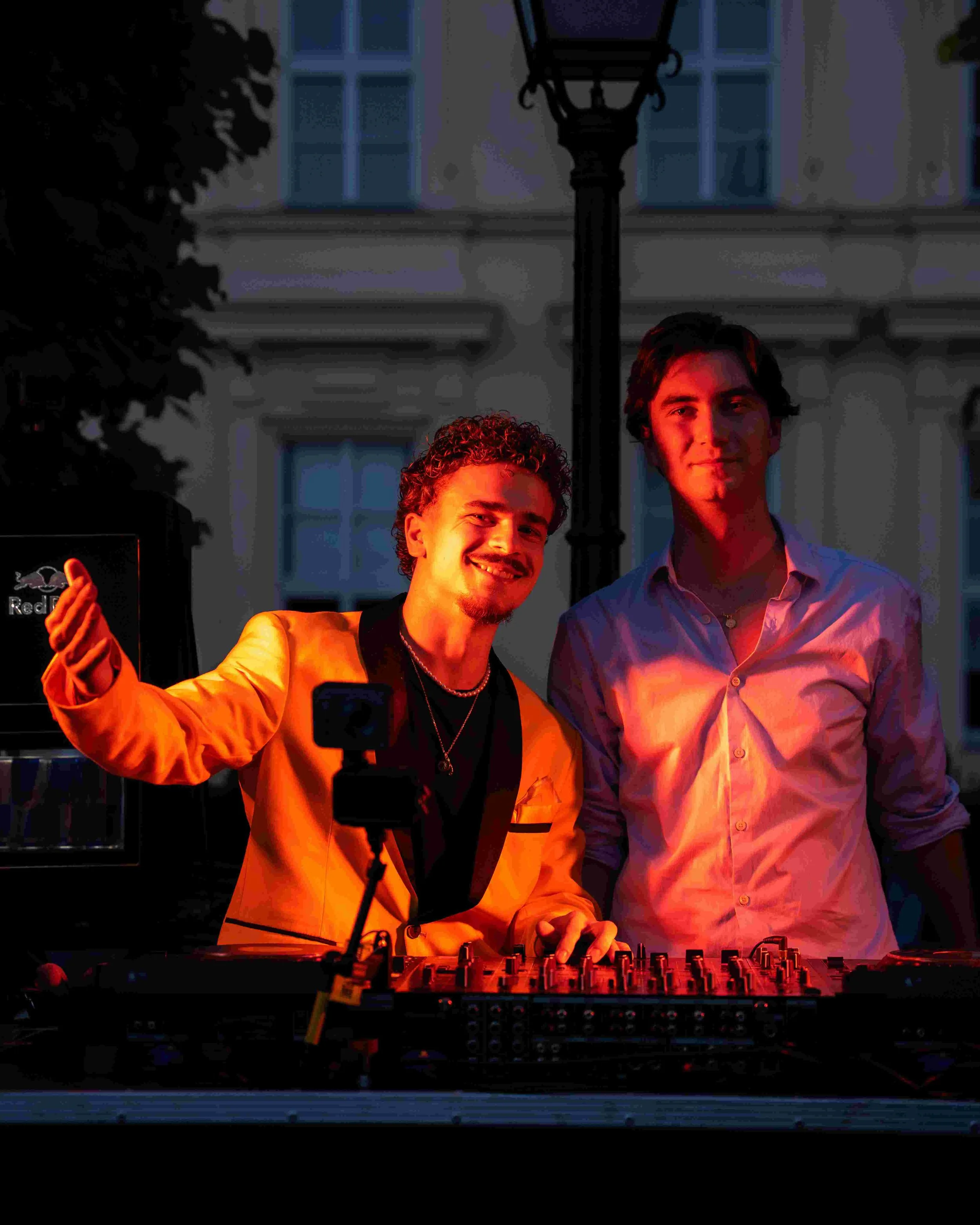 Two young men posed for a photo outdoors at night, one wearing an orange blazer and the other a light-colored shirt, standing behind a DJ mixer, with a streetlamp and building windows illuminated in the background.