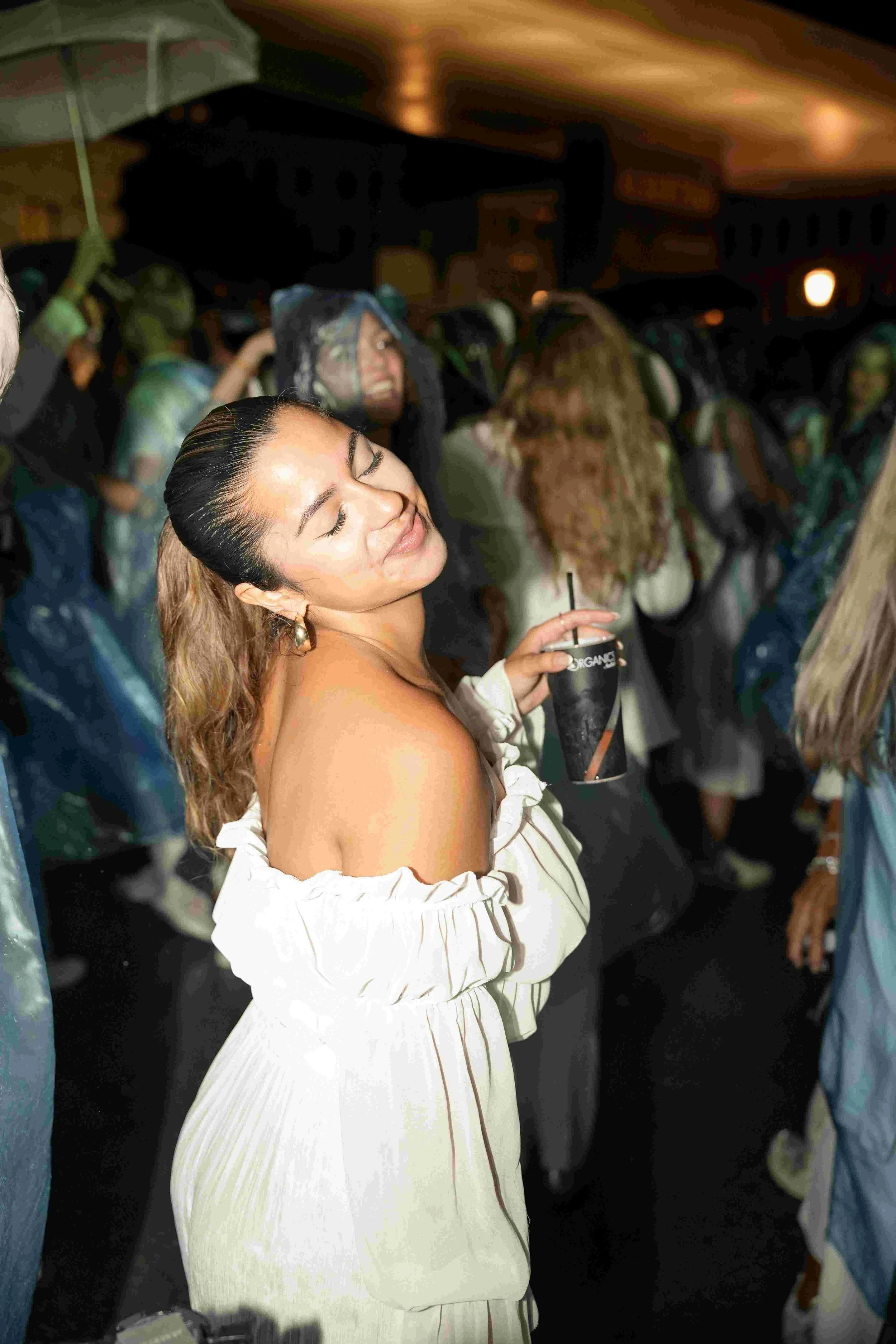 A woman with long, wavy hair tied back, wearing an off-the-shoulder white dress with ruffles, smiling with eyes closed, holding a drink with a straw in a black cup, at a crowded indoor event.