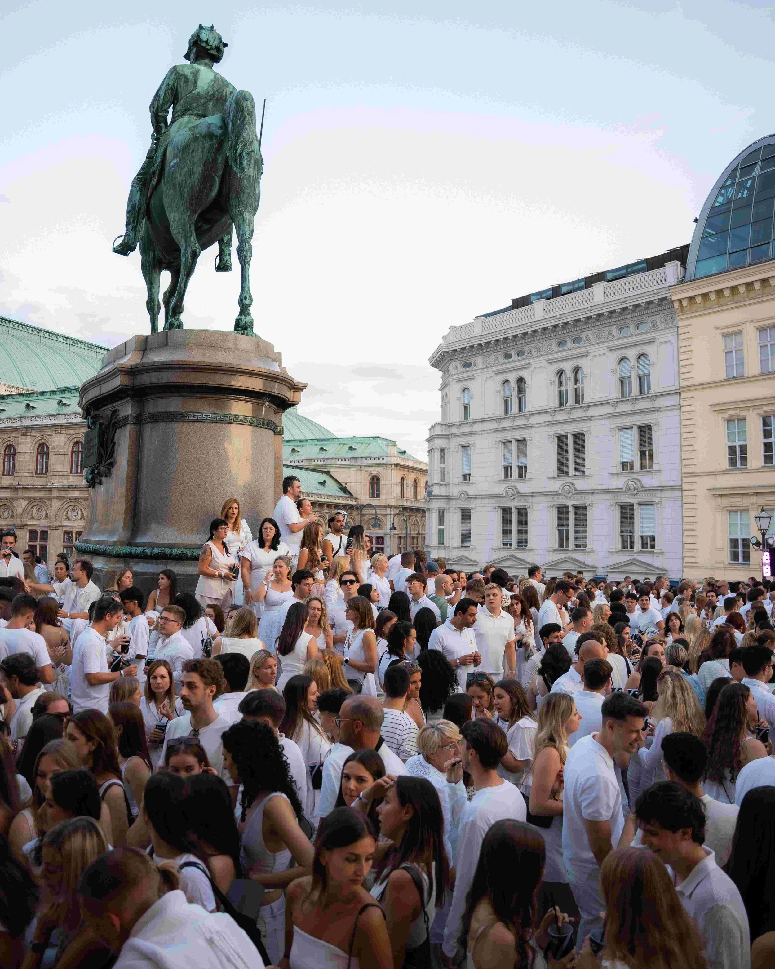 A large crowd of people gathered in front of a historic building with a prominent equestrian statue on a pedestal. Most of the crowd is wearing white. The sky is clear, and the background features a mix of classical and modern architecture.