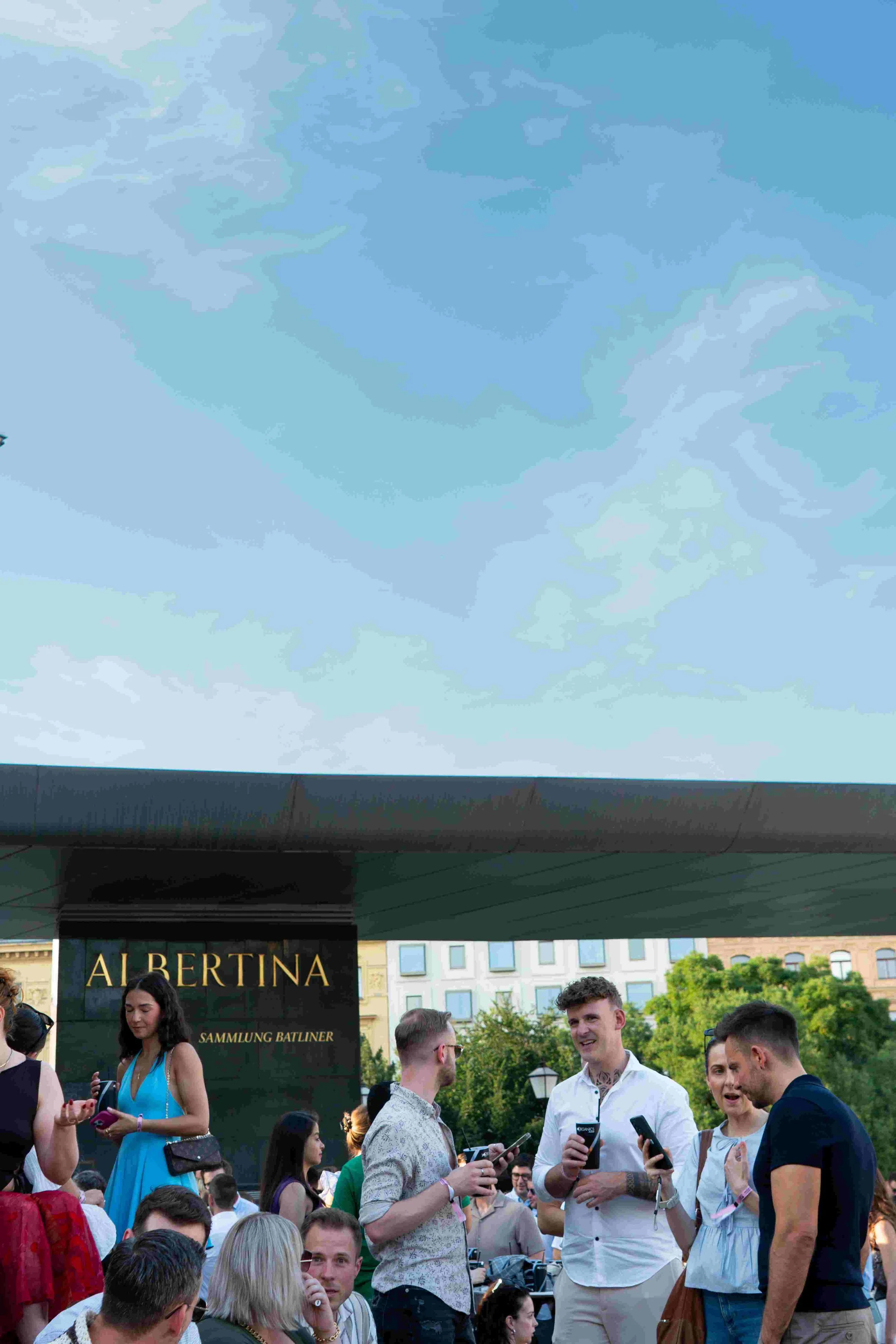 A group of people at an outdoor event surrounding a man in a white shirt talking on his phone, with a black sign reading "AI BERTINA" and a cityscape with buildings and trees in the background.