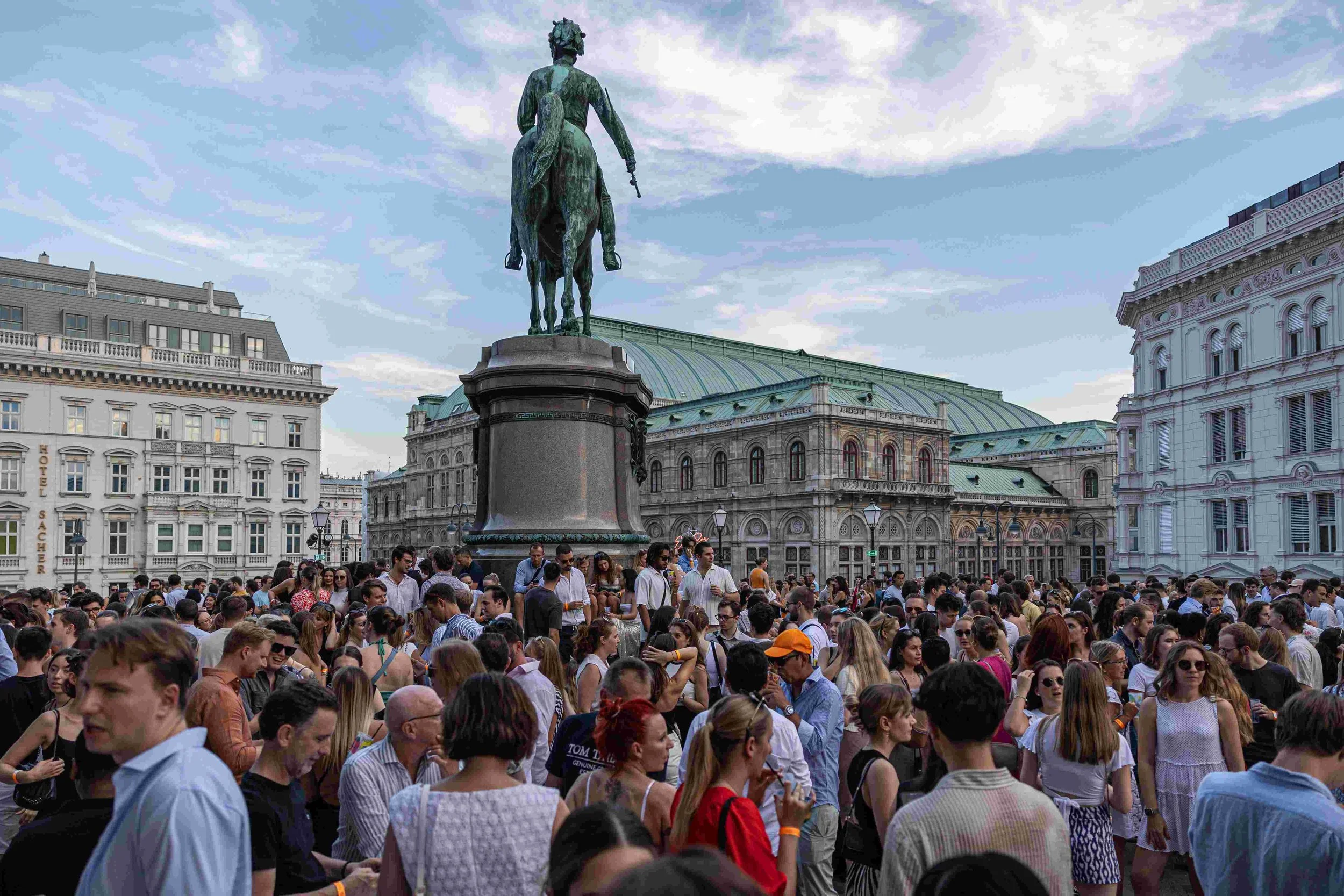 A large crowd of people gathered outdoors in front of an equestrian statue of a man with a crown or helmet, situated on a city square with historic buildings and a partly cloudy sky overhead.