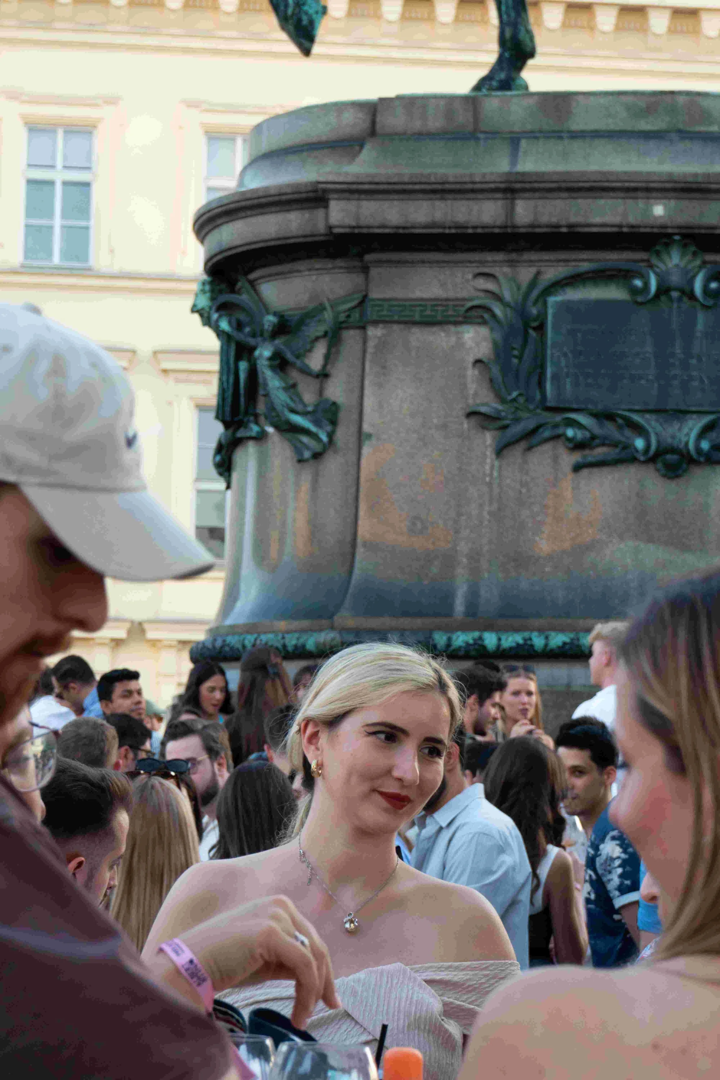 A crowd of people gathered outdoors in front of a historical building with windows. In the center, a young woman with blonde hair and earrings is looking slightly down and smiling. The image shows a variety of facial expressions and interactions amon