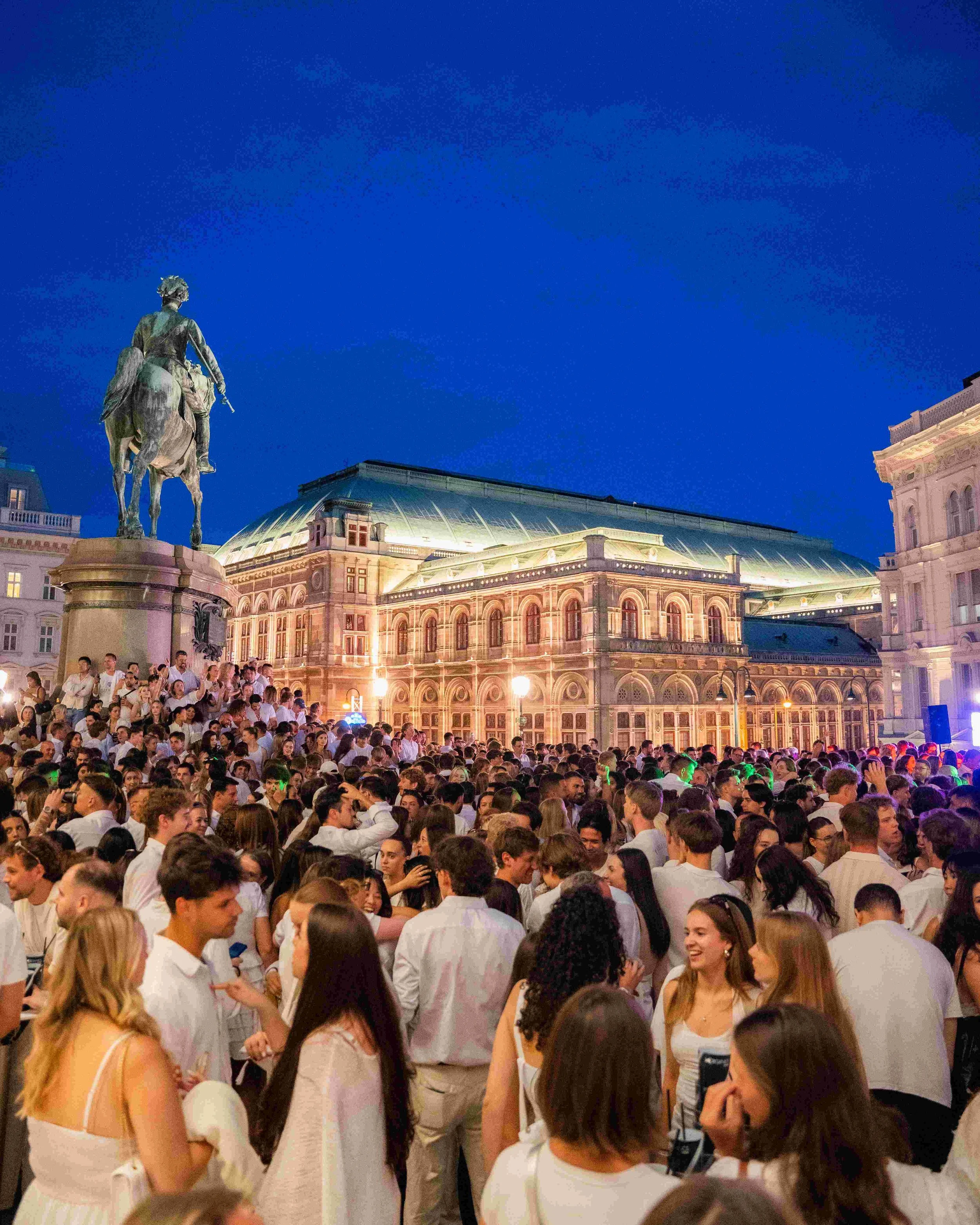 Large crowd gathered outdoors at night in front of historic building with domed roof and statue of a man on horseback.