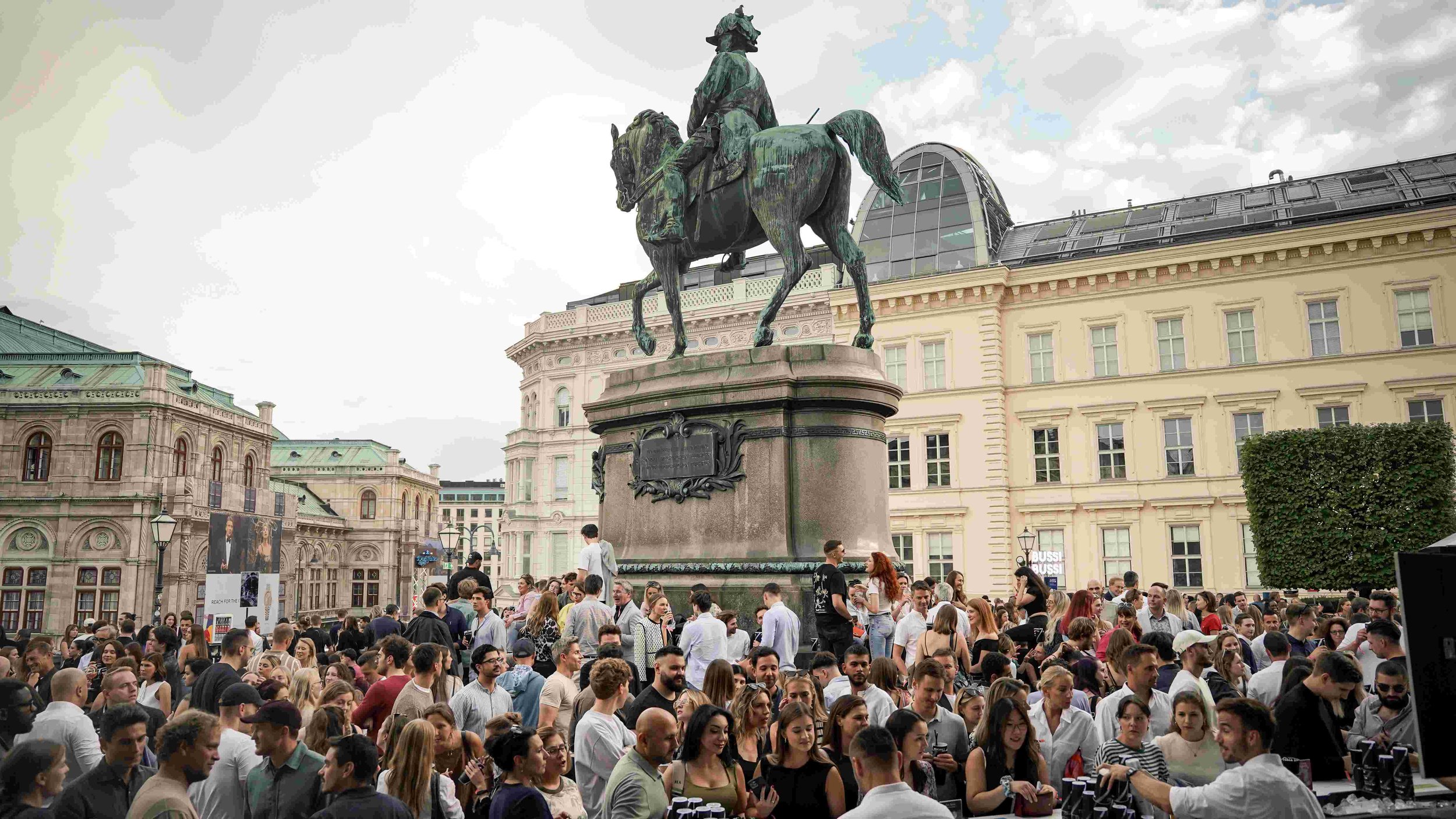 A busy crowd of people gathered around a large bronze equestrian statue in an urban square surrounded by historic buildings.