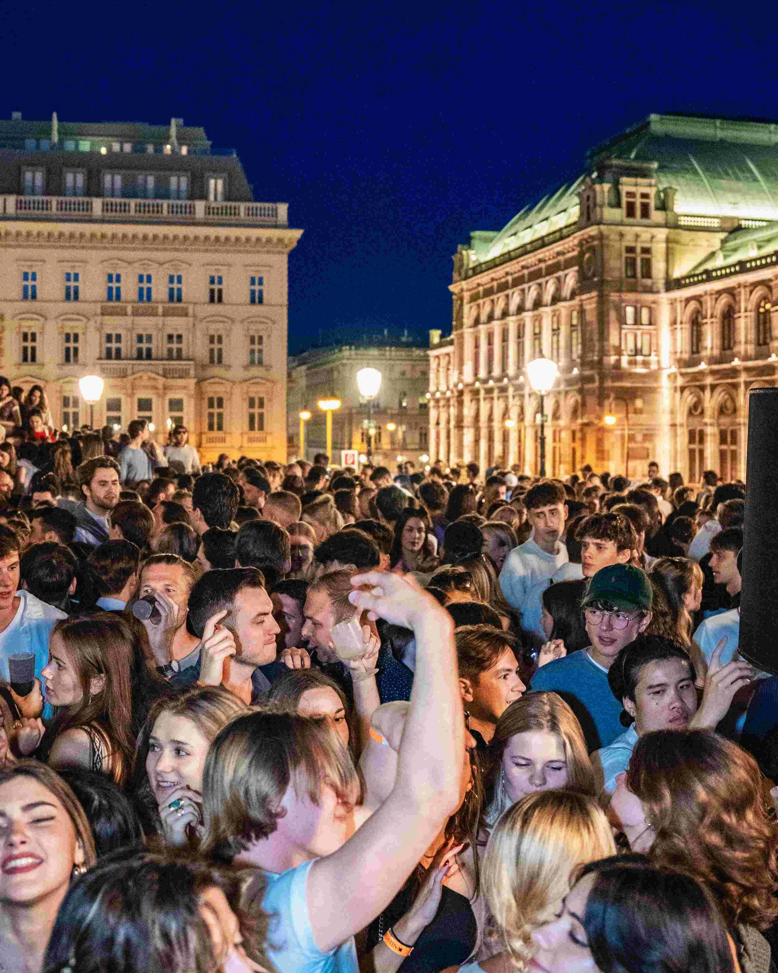 A large crowd of young people gathered in an outdoor urban setting at night, with historic buildings and street lamps illuminated in the background.