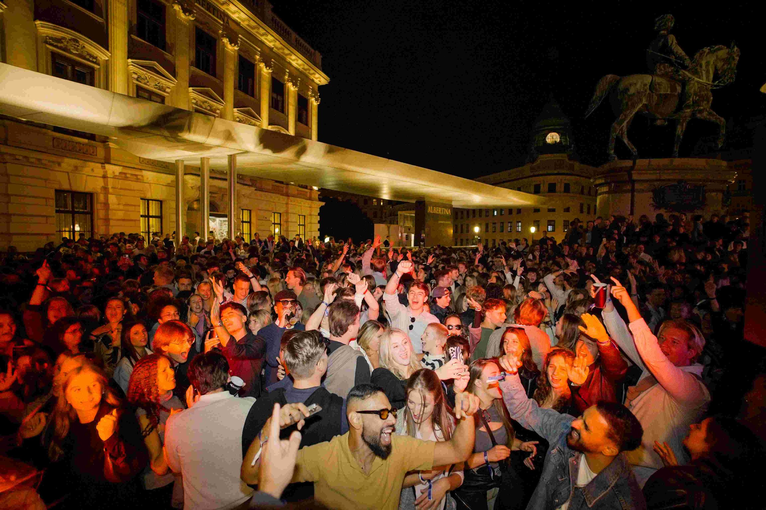A large crowd of people gathered outside a historic building at night, some dancing and taking photos. The building has ornate architecture and a long, illuminated canopy. An equestrian statue is visible in the background.