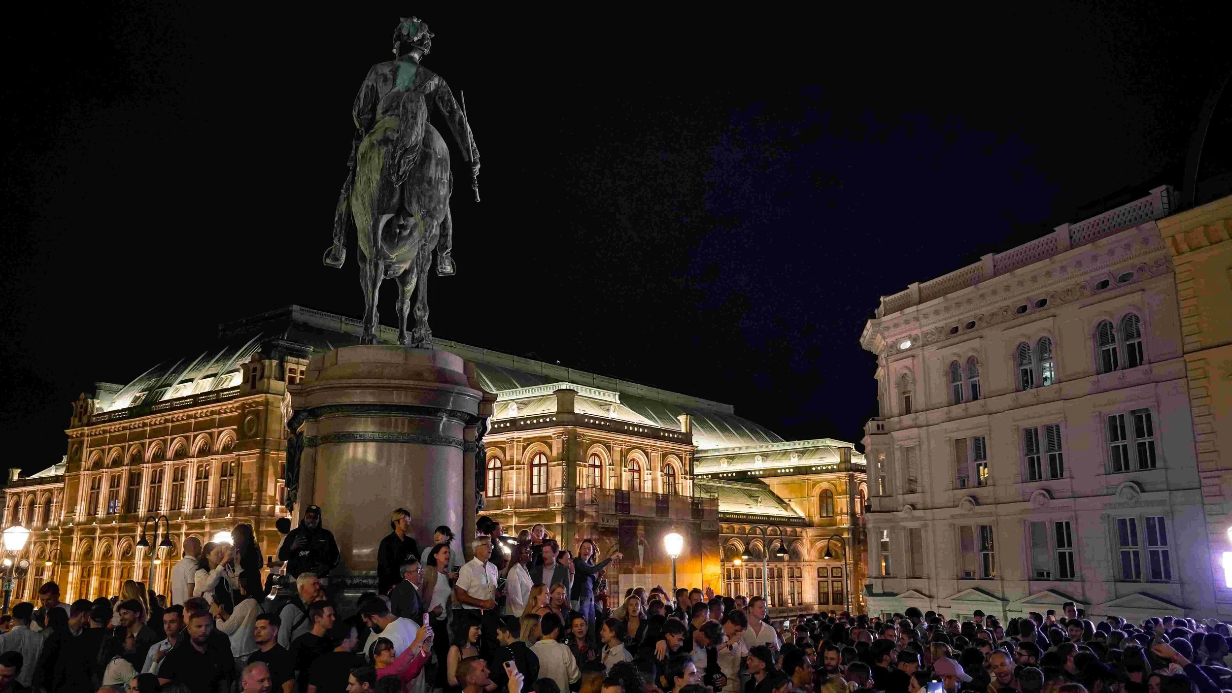 Night view of a crowded outdoor event in front of an illuminated historic building with a statue of a man on horseback on a pedestal.