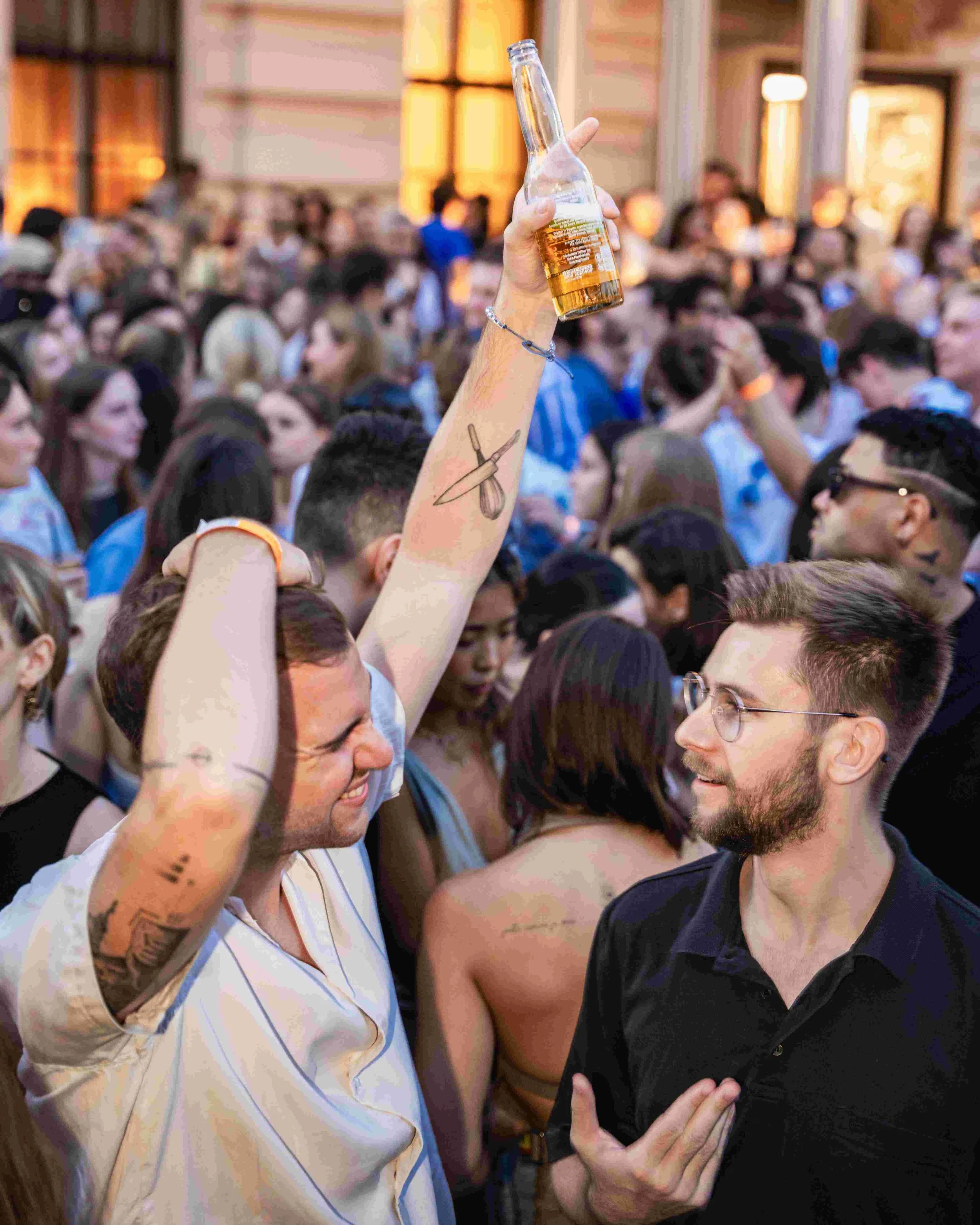 A crowd of people at a social gathering, with one person holding up a beer bottle in the foreground. The scene is lively, with individuals engaging and smiling, set indoors with warm lighting.