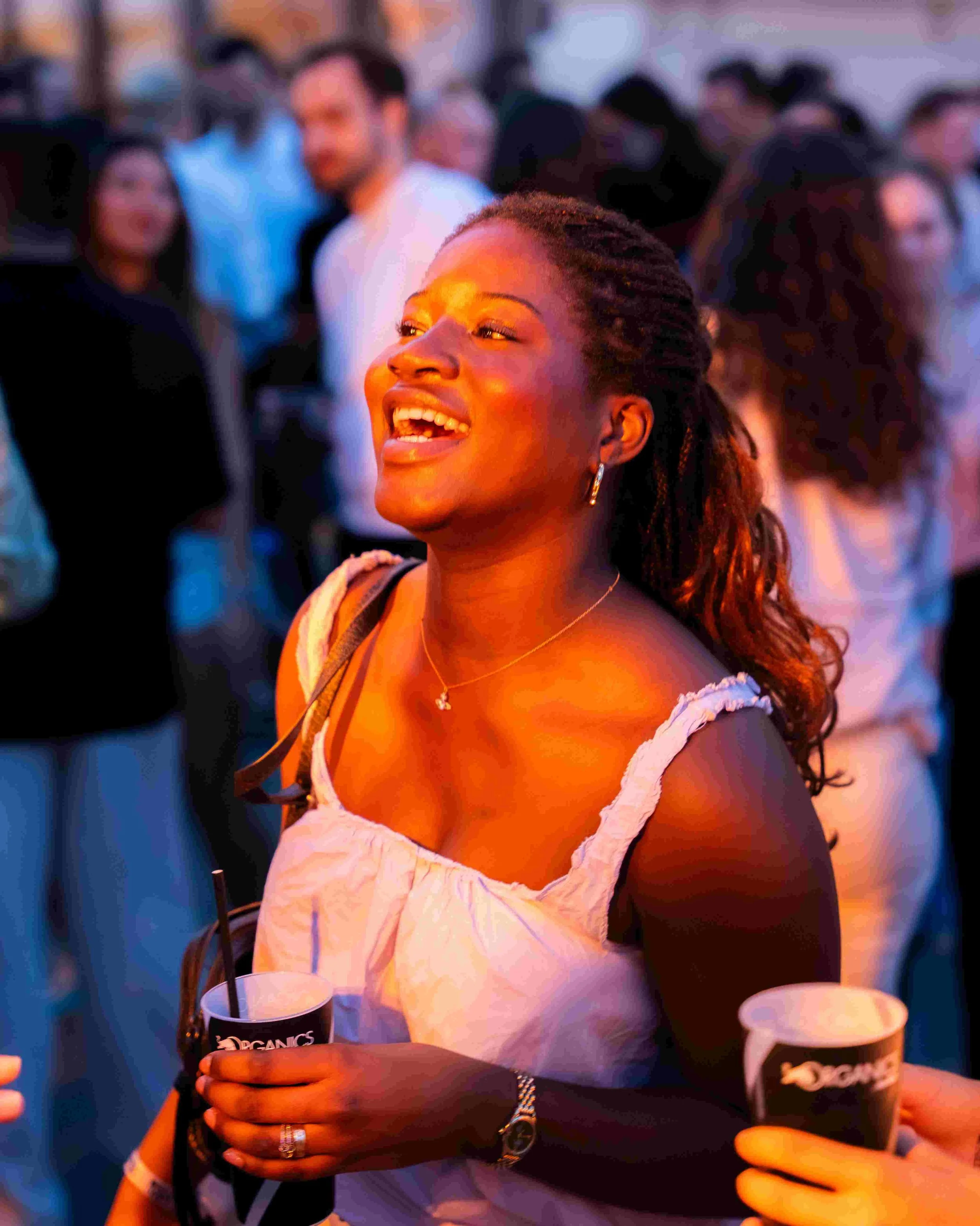 A woman smiling at a social gathering, holding a black cup with the logo 'ORGANICS' in her right hand. People in the background are blurred out, indicating a lively atmosphere. The woman has dark braided hair, gold earrings, and is wearing a white sl