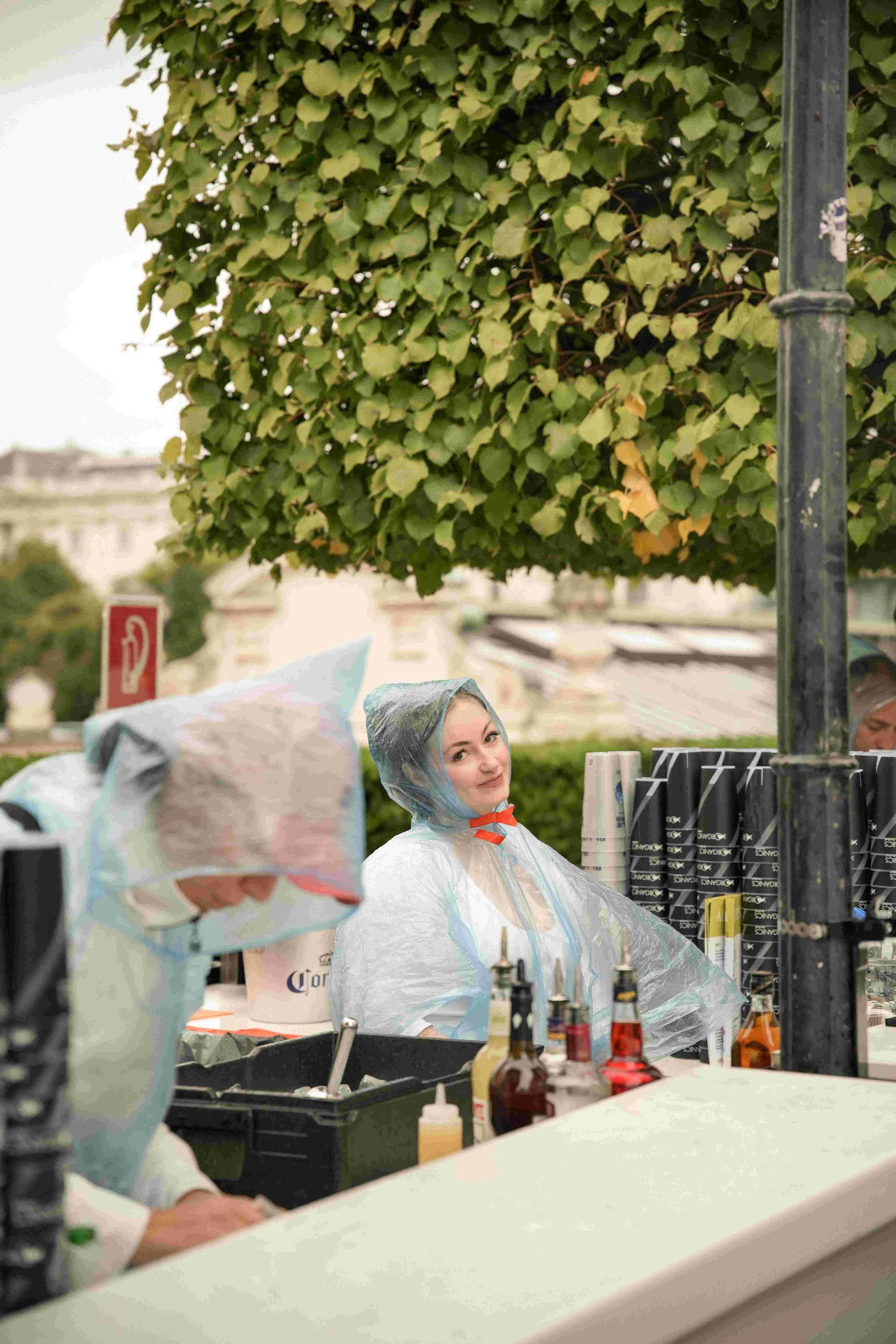 People wearing plastic rain ponchos at an outdoor event, with a table of condiments in the foreground and a lush green tree overhead.