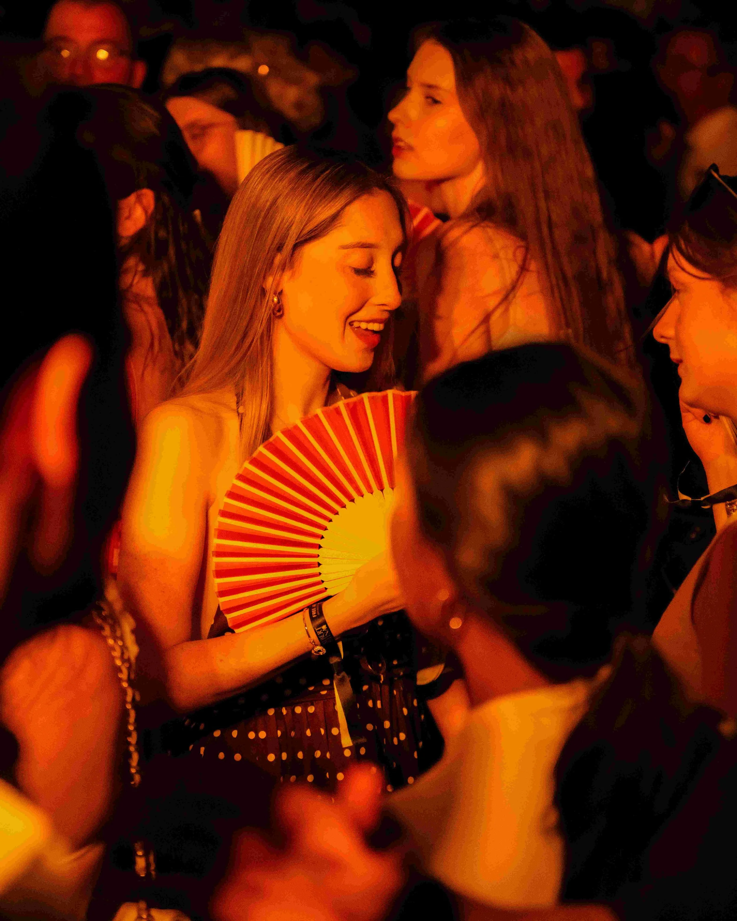 Two women in a dimly lit setting, one smiling and holding a red and yellow striped fan, the other with long hair looking to the side, surrounded by other people.
